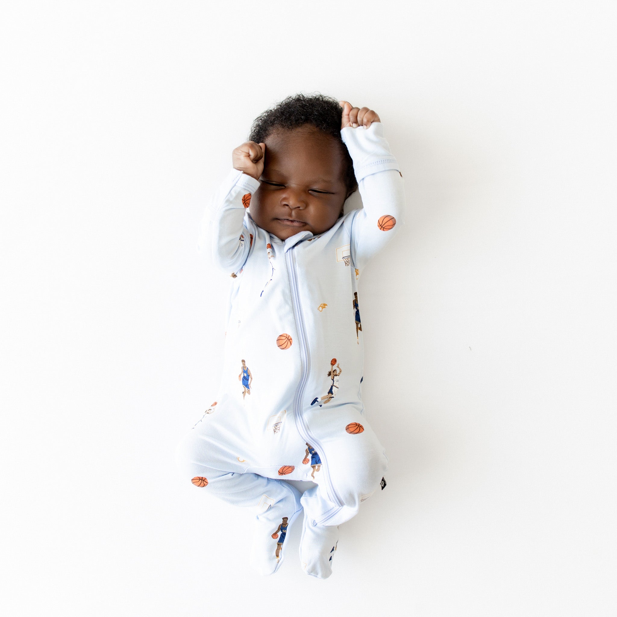 Sleeping infant wearing the Zippered Footie in Basketball with their hands resting on their head