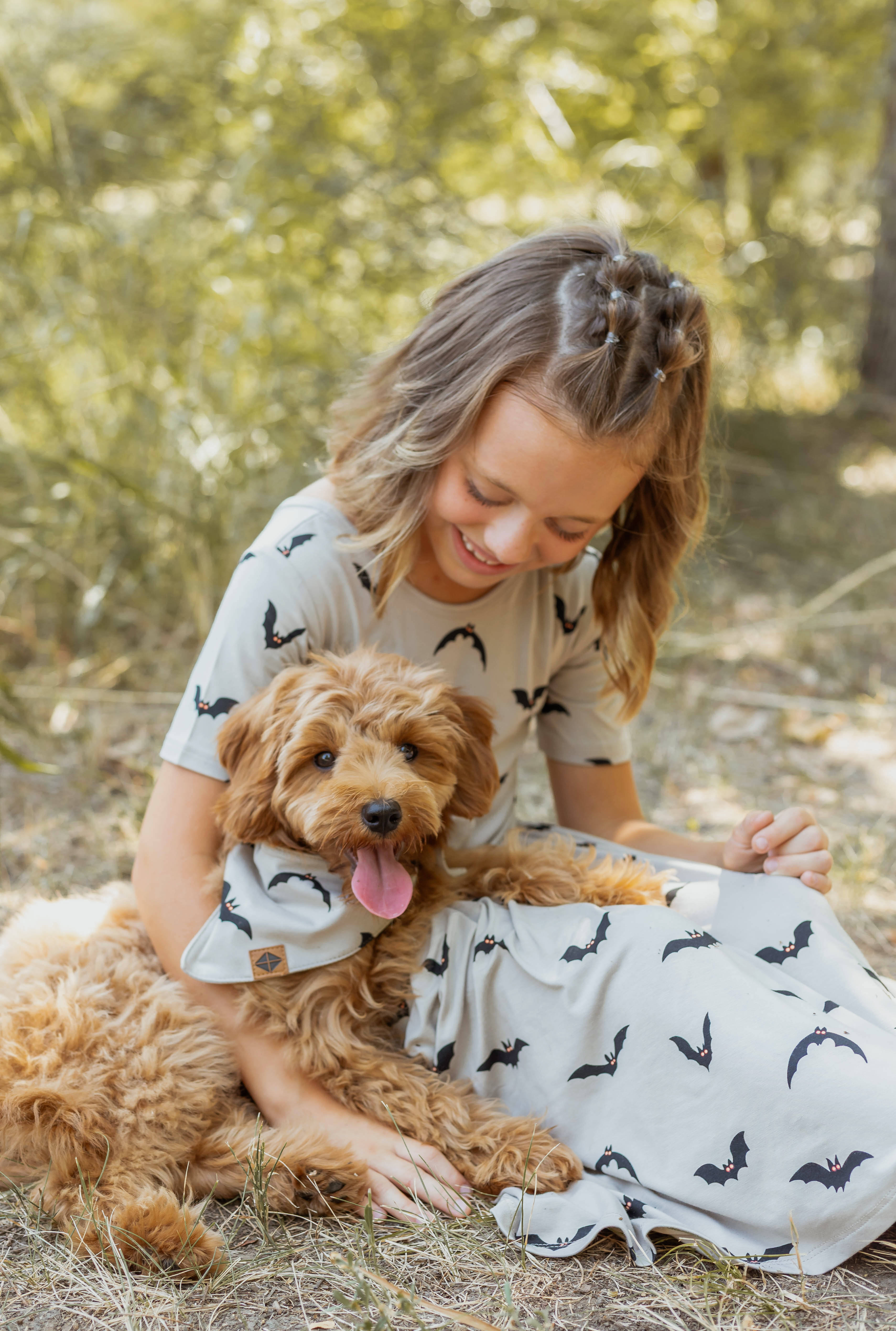 A girl sitting with a Brown dog in Dog Bandana in Bats print wearing a similar print twirl dress on grass outdoors