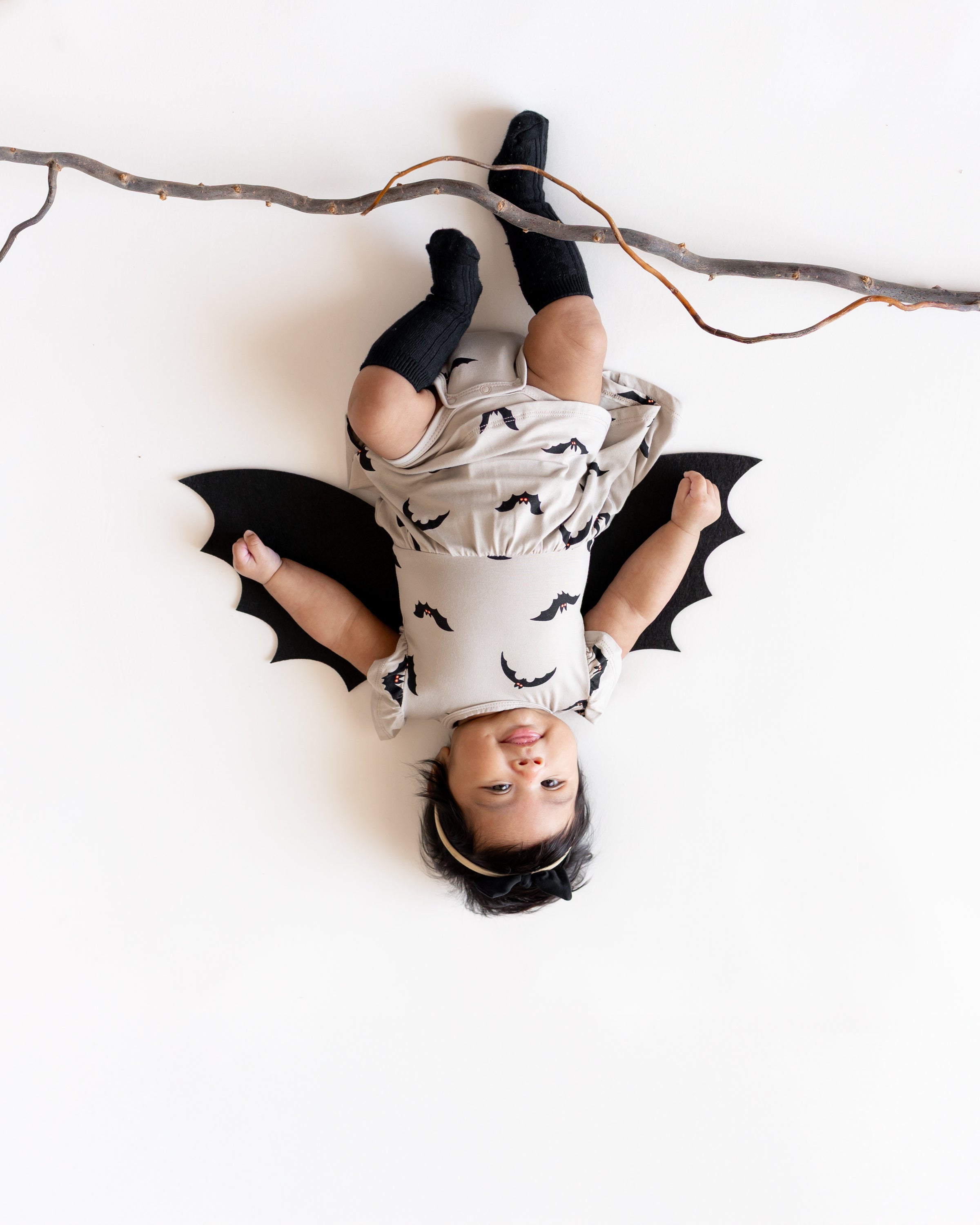 Baby posing as a bat hanging upside down from a branch wearing Twirl Bodysuit Dress in Bats print with bat wings and black socks on a white background
