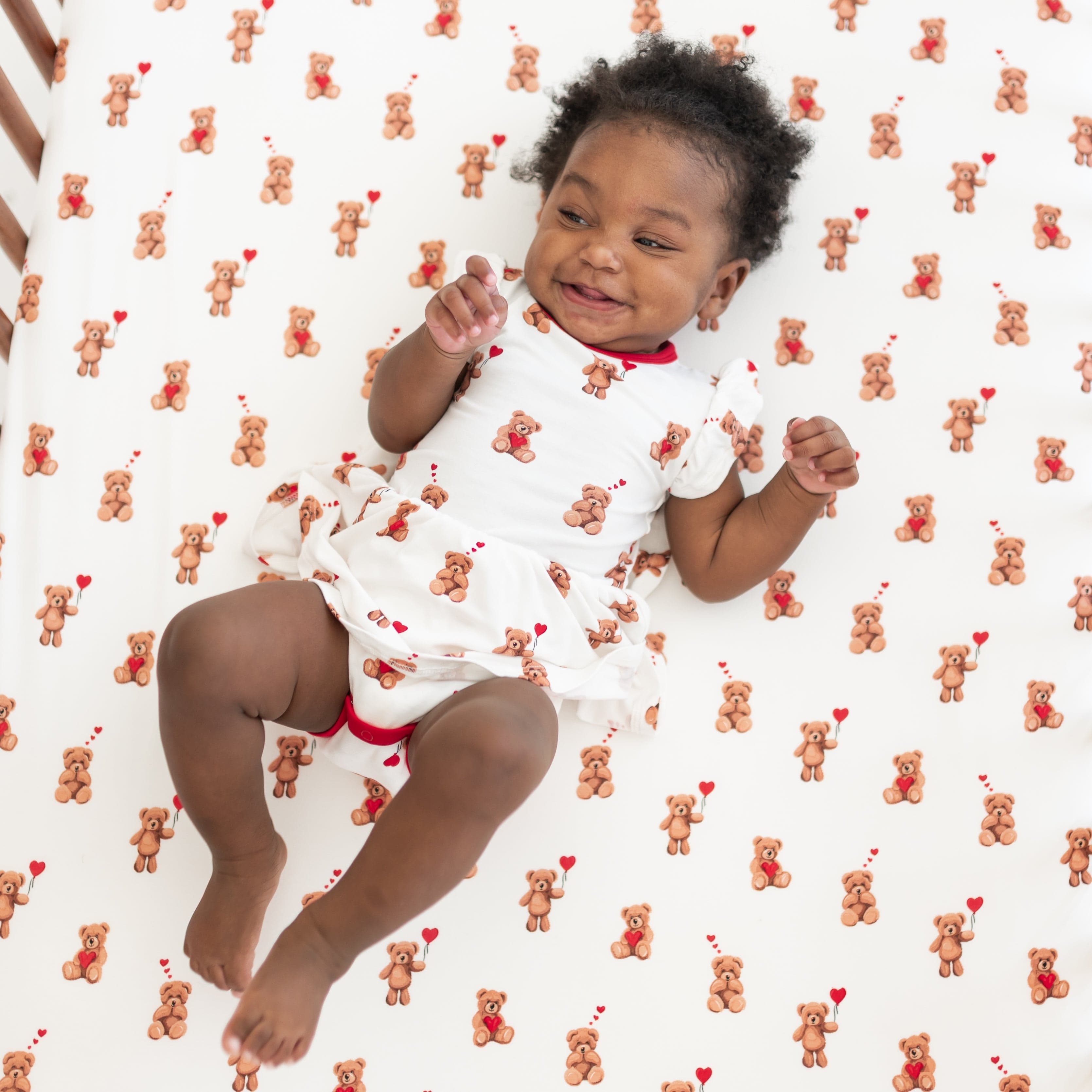 Smiling toddler laying in a crib on a Crib Sheet in Bear Hearts wearing a matching twirl bodysuit dress