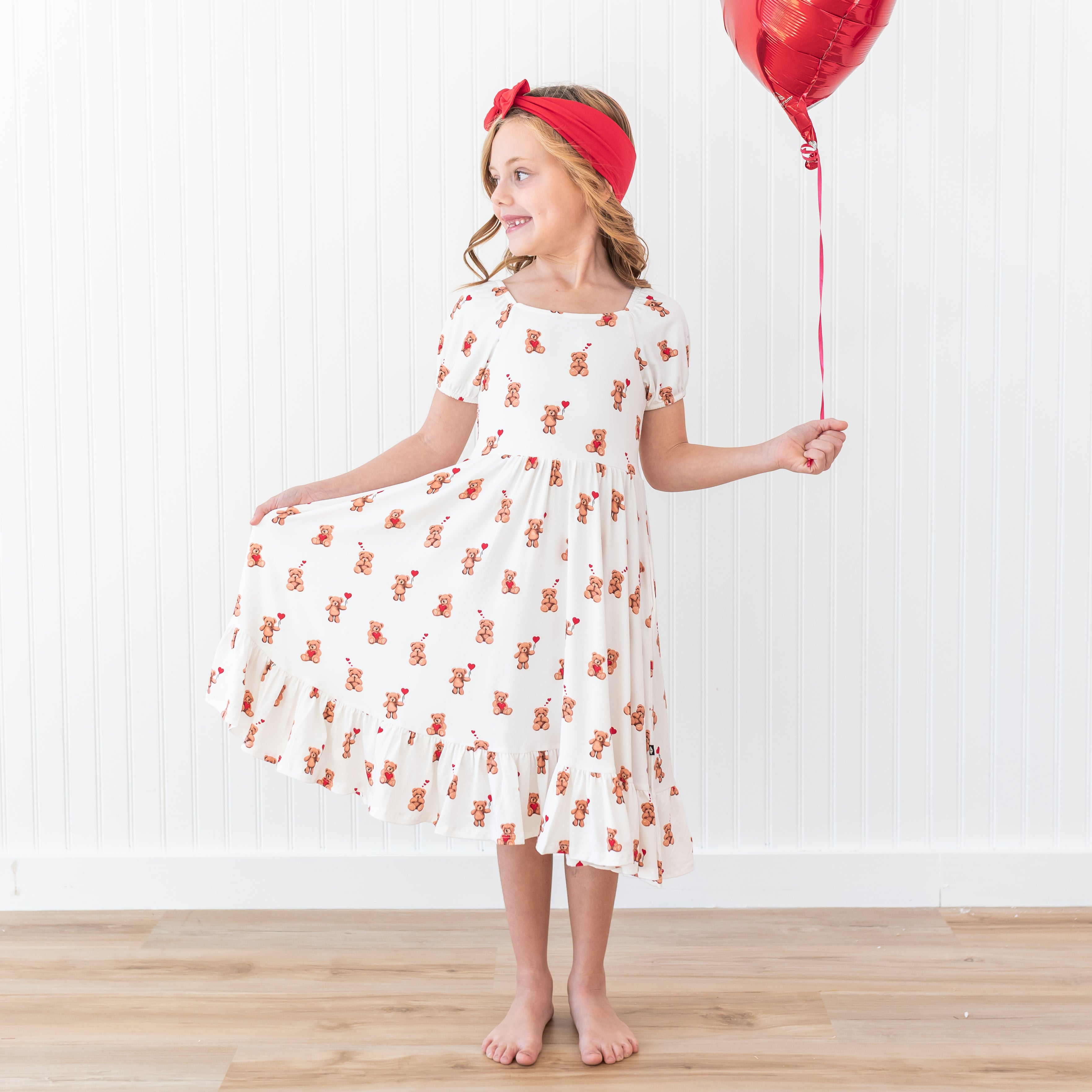 Young girl wearing the Puff Sleeve Twirl Dress in Bear Hearts with a Cardinal Knotted bow headband holding a red heart shaped balloon with one hand and the skirt of her dress in the other