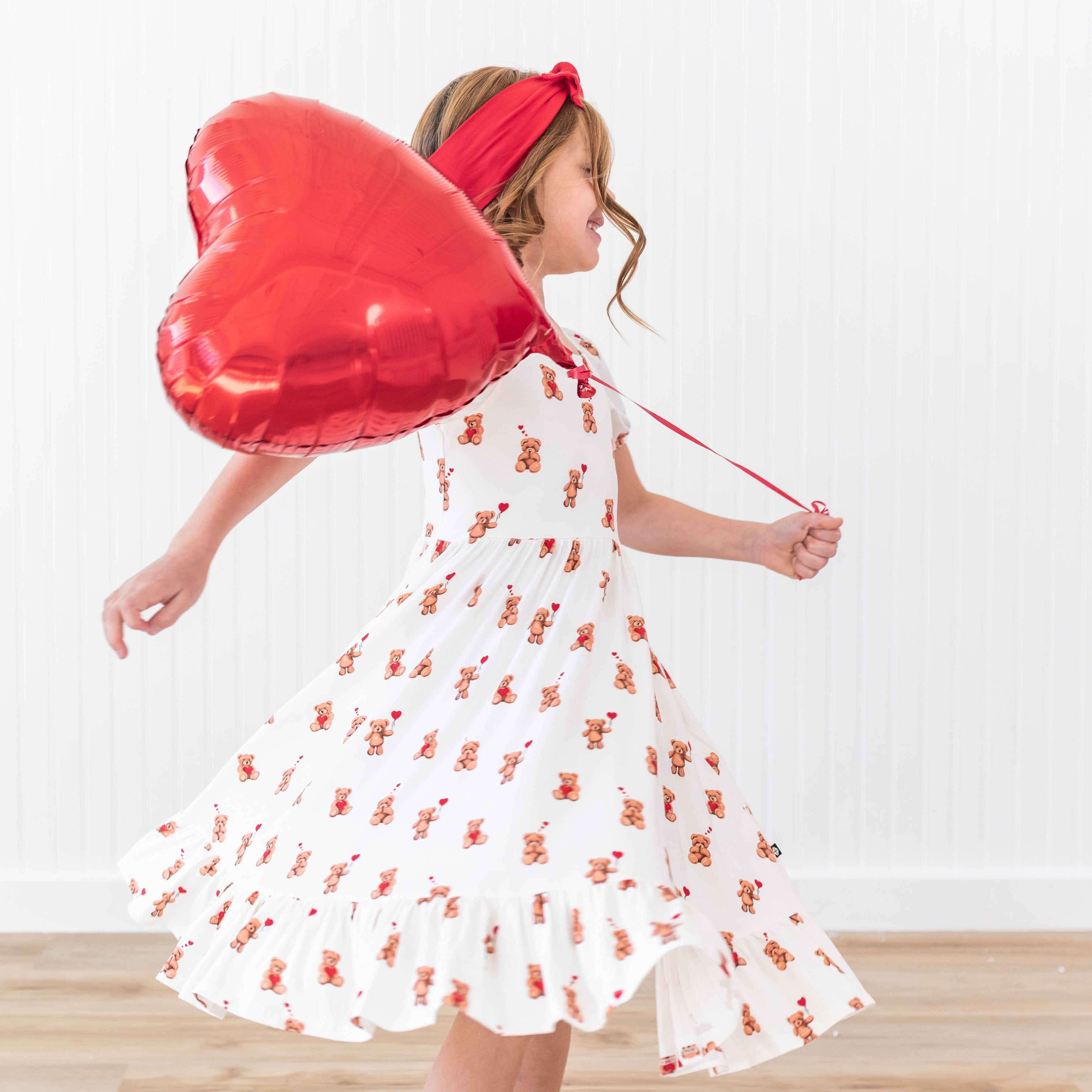 Young girl twirling in her flowy Puff Sleeve Twirl Dress in Bear Hearts holding a red heart balloon