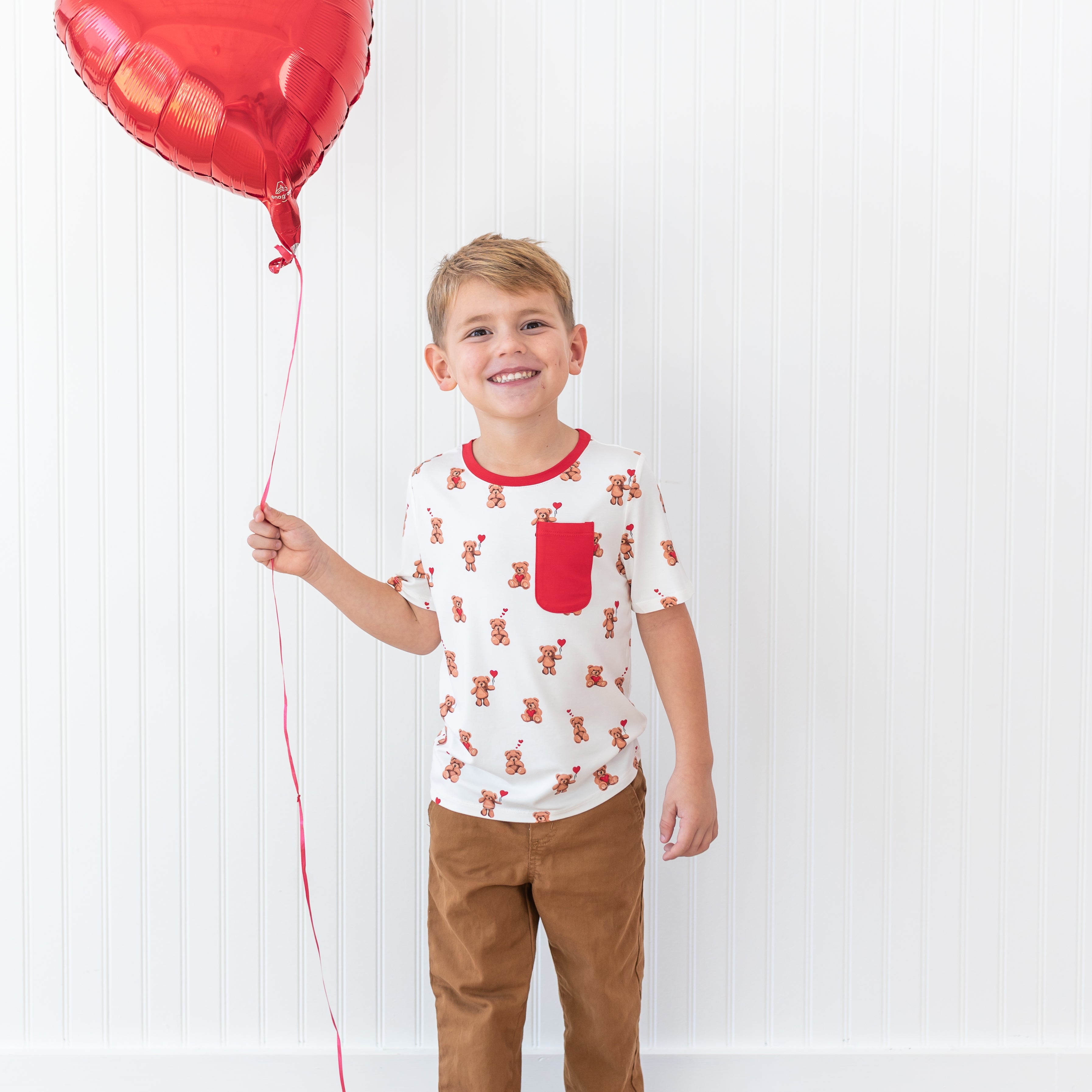 Smiling boy standing in front of a white paneled wall holding a red heart shaped balloon wearing the soft and light Toddler Crew Neck Tee in Bear Hearts