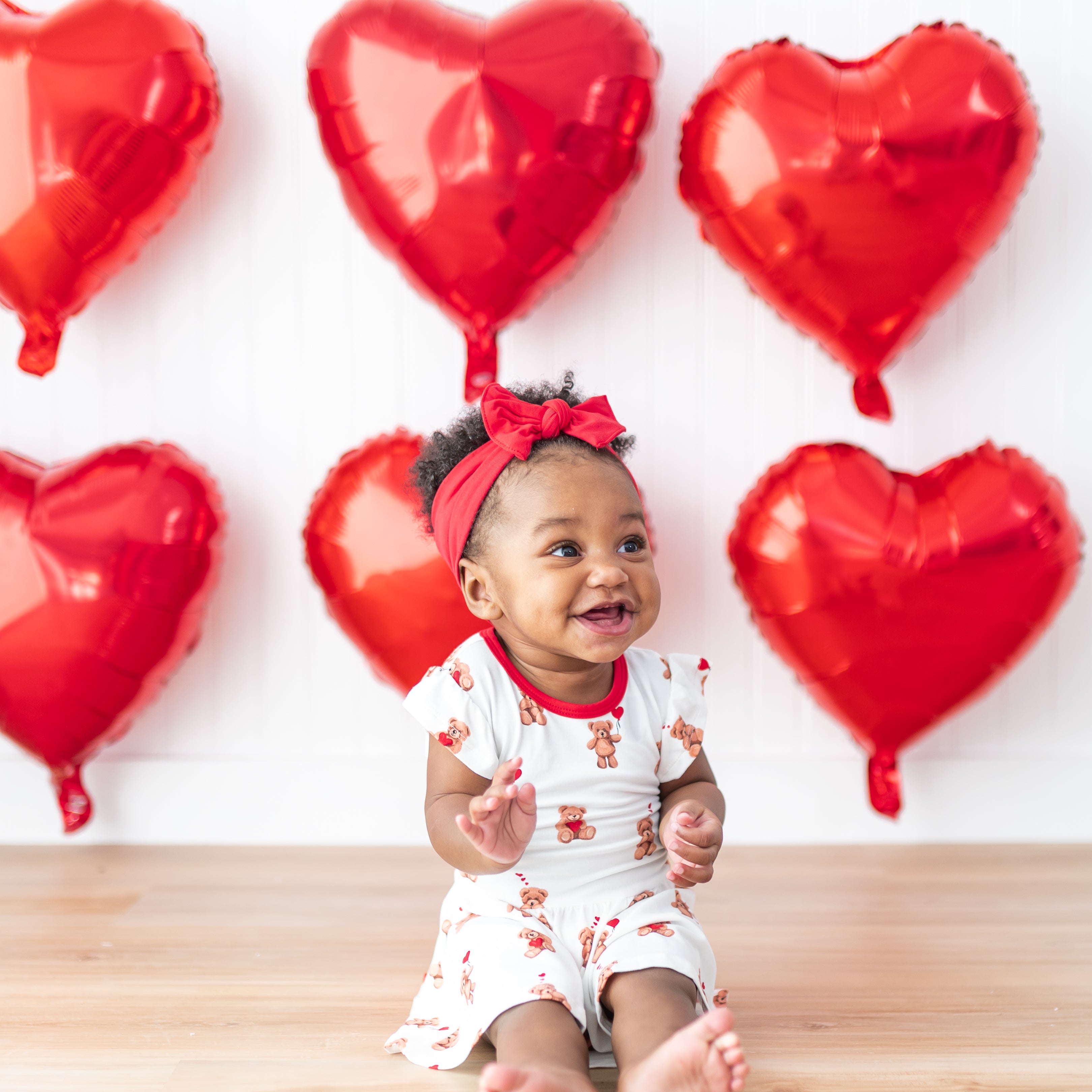 Toddler sitting on the floor in front of red heart balloons on a white wall wearing the Twirl Bodysuit Dress in Bear Hearts and cardinal knotted bow headband