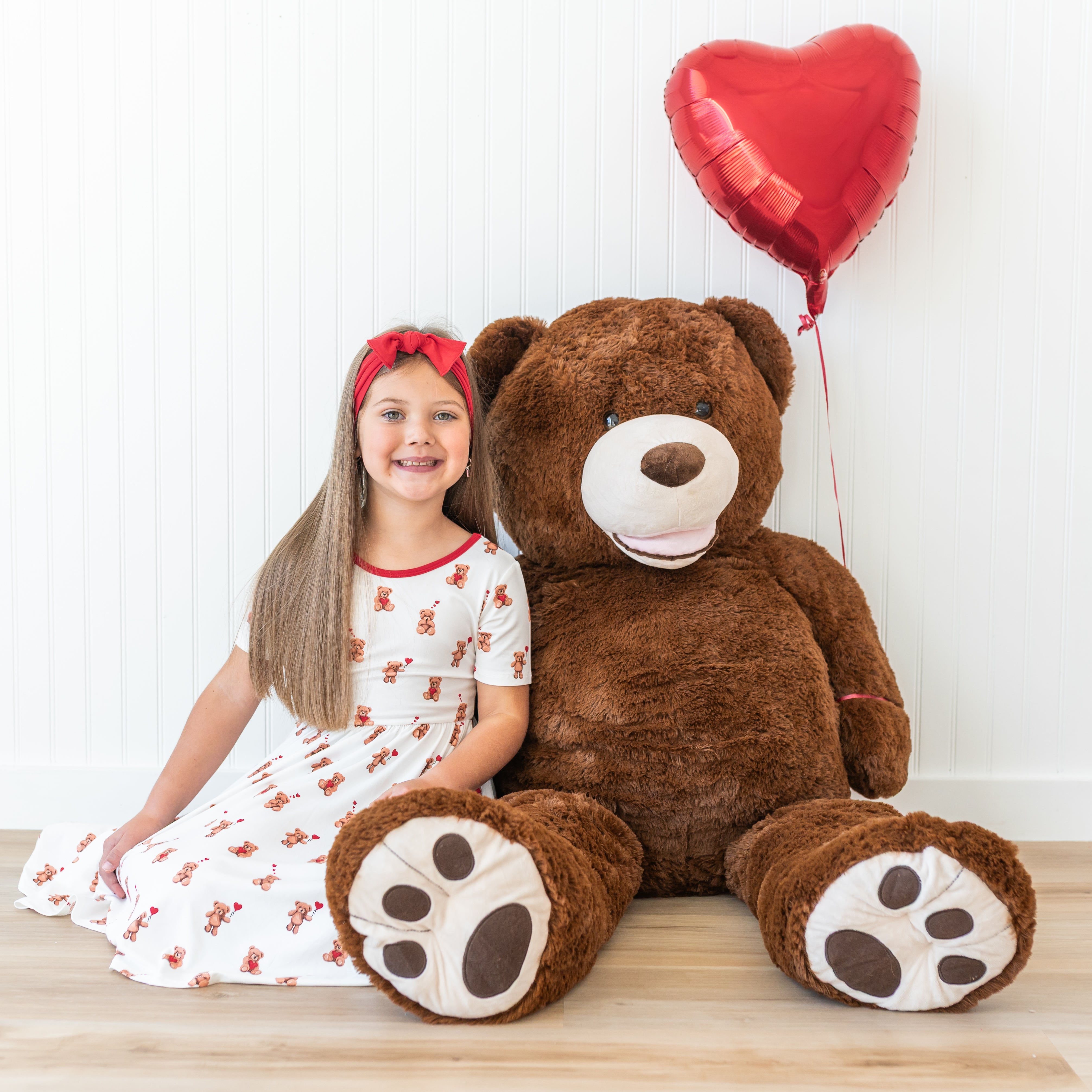 Young girl wearing the Twirl Dress in Bear Hearts sitting on the floor beside a large stuffed bear with a red heard balloon tied to its arm