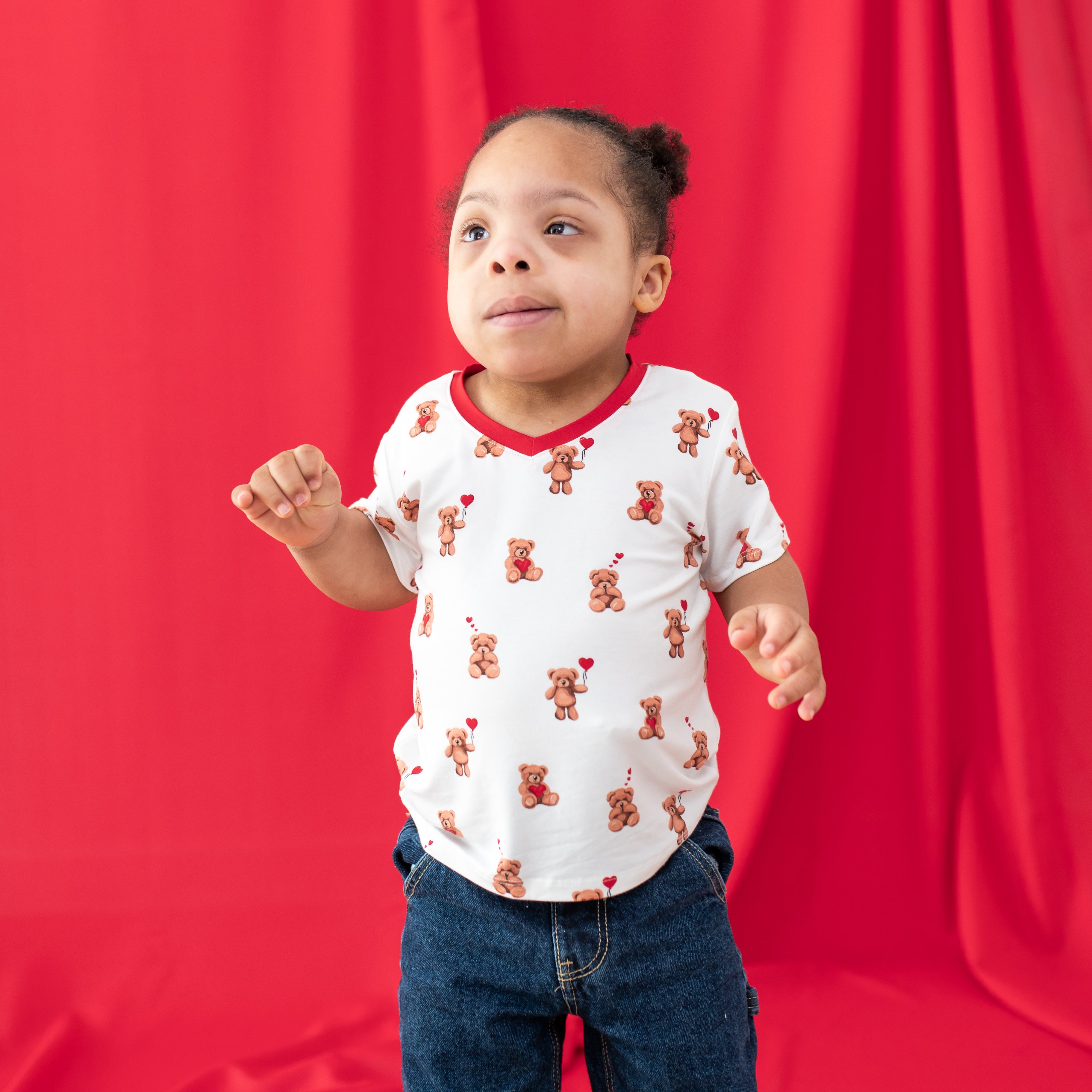 young girl standing in front of a curtain wearing the Toddler V-Neck Tee in Bear Hearts paired with medium wash jeans