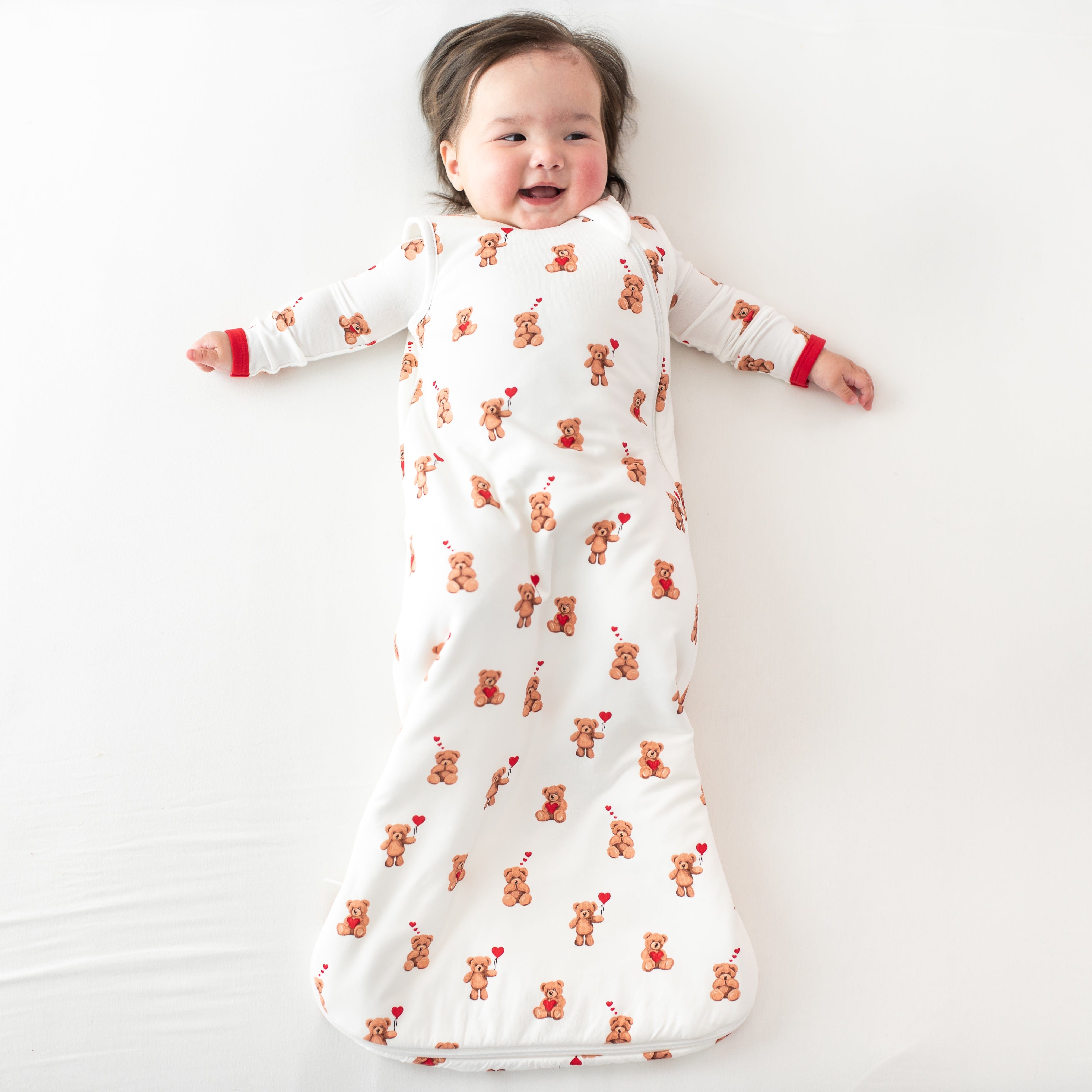 Smiling infant laying on a light neutral blanket wearing the Sleep Bag in Bear Hearts 1.0 with a matching romper on underneath