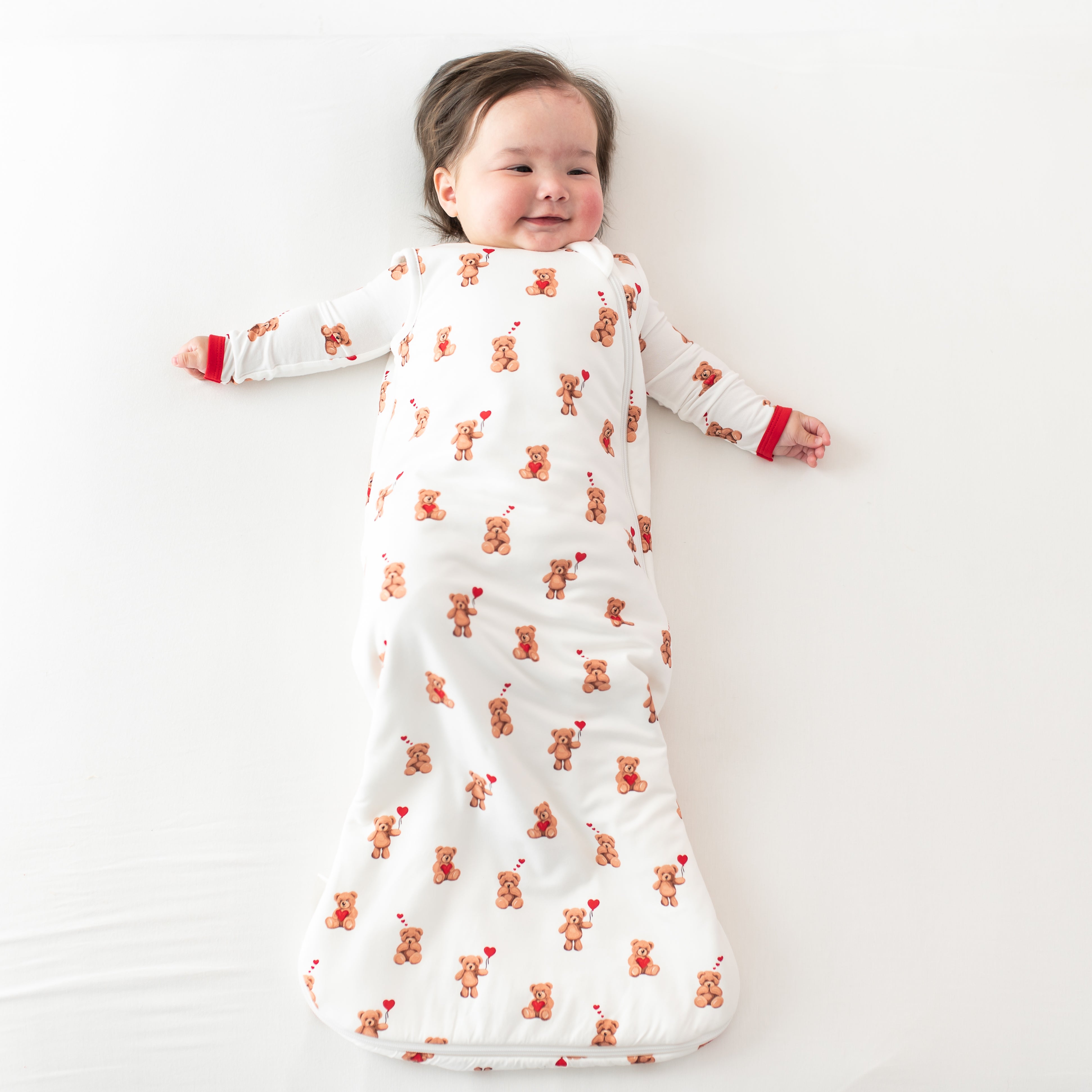Smiling infant laying on a light colored blanket wearing the Sleep Bag in Bear Hearts 1.0 with matching romper on underneath