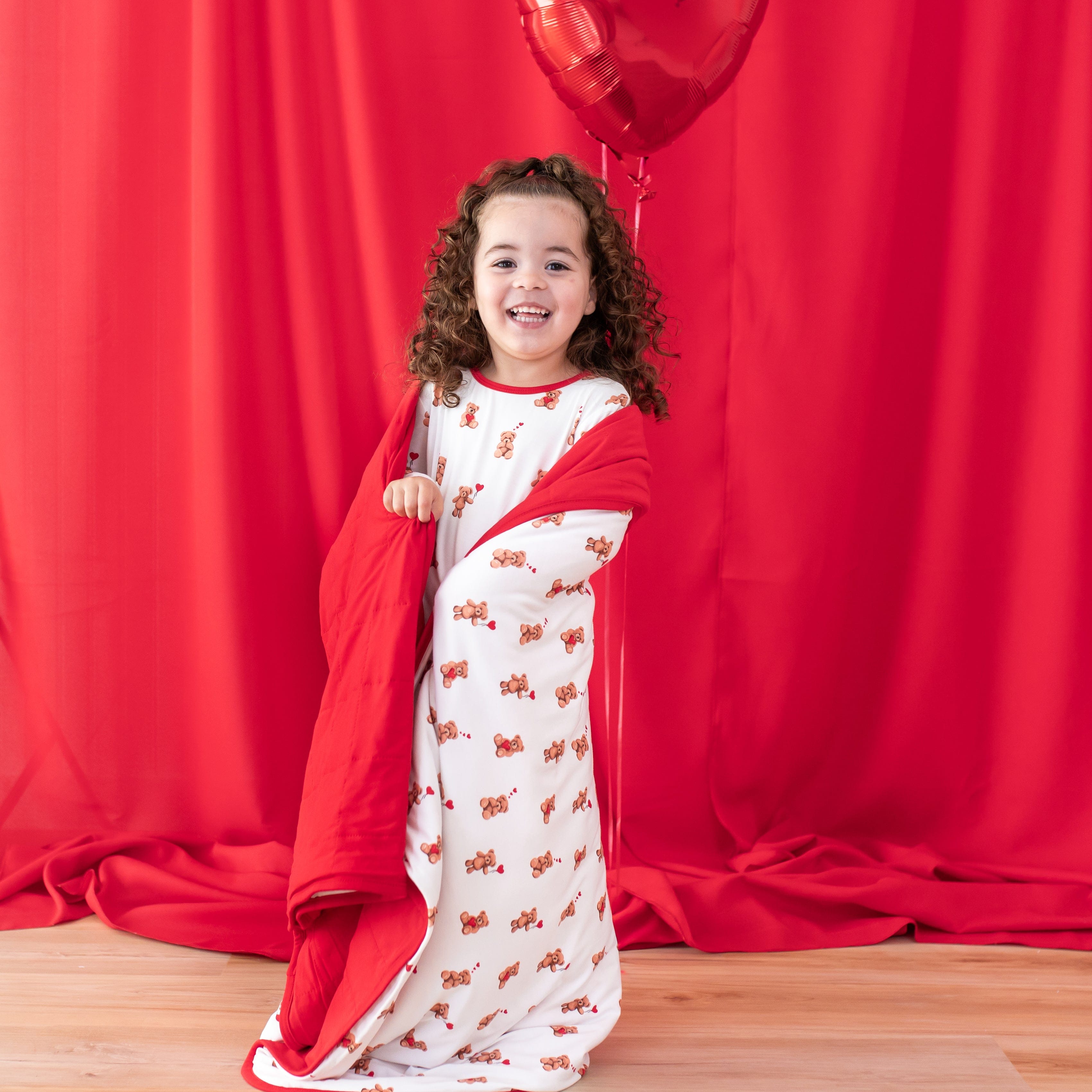 Smiling girl standing in front of a red curtain with the Toddler Blanket in Bear Hearts 1.0 wrapped around her