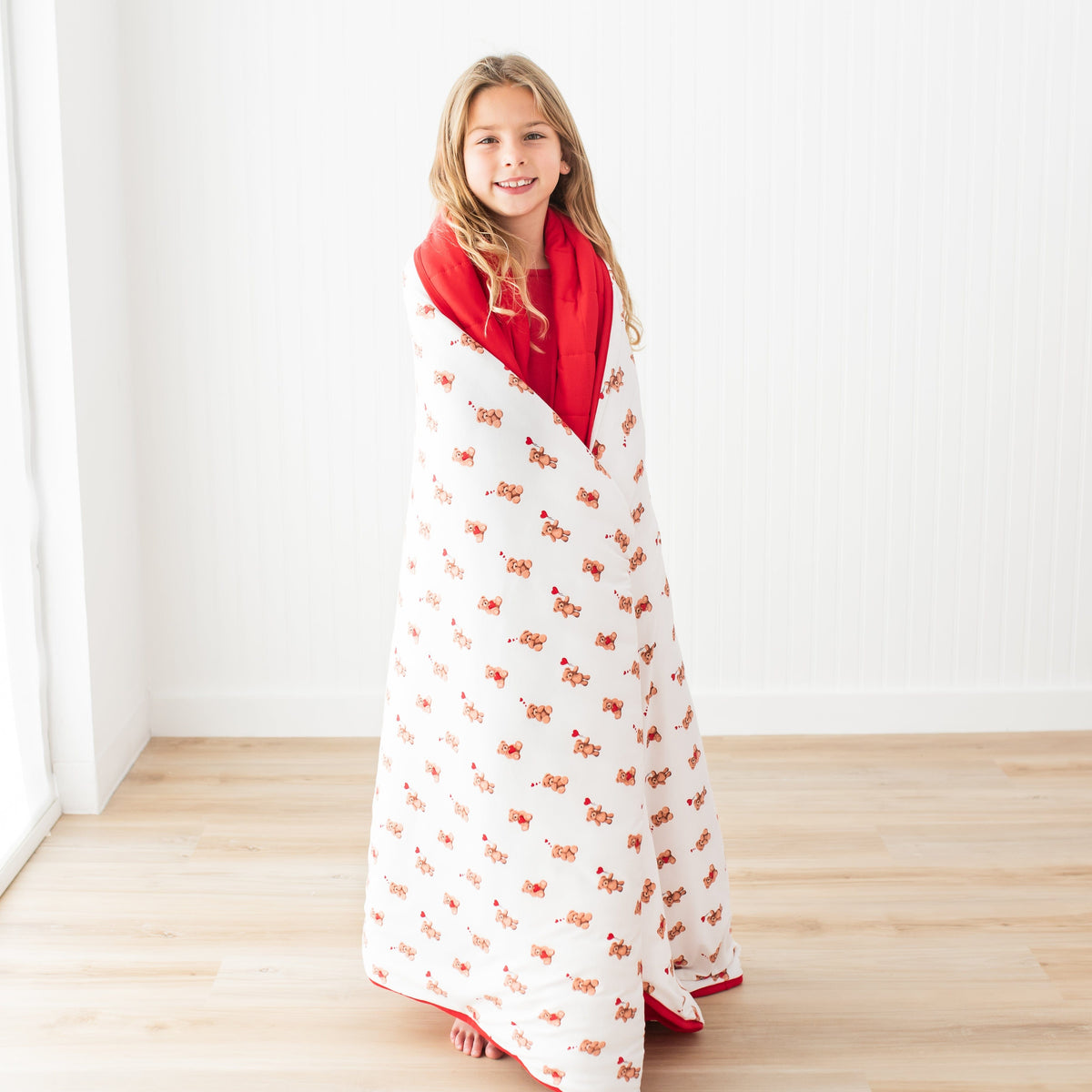 Young girl standing in front of a white paneled wall wearing the Youth Blanket in Bear Hearts 2.5 wrapped around her shoulders