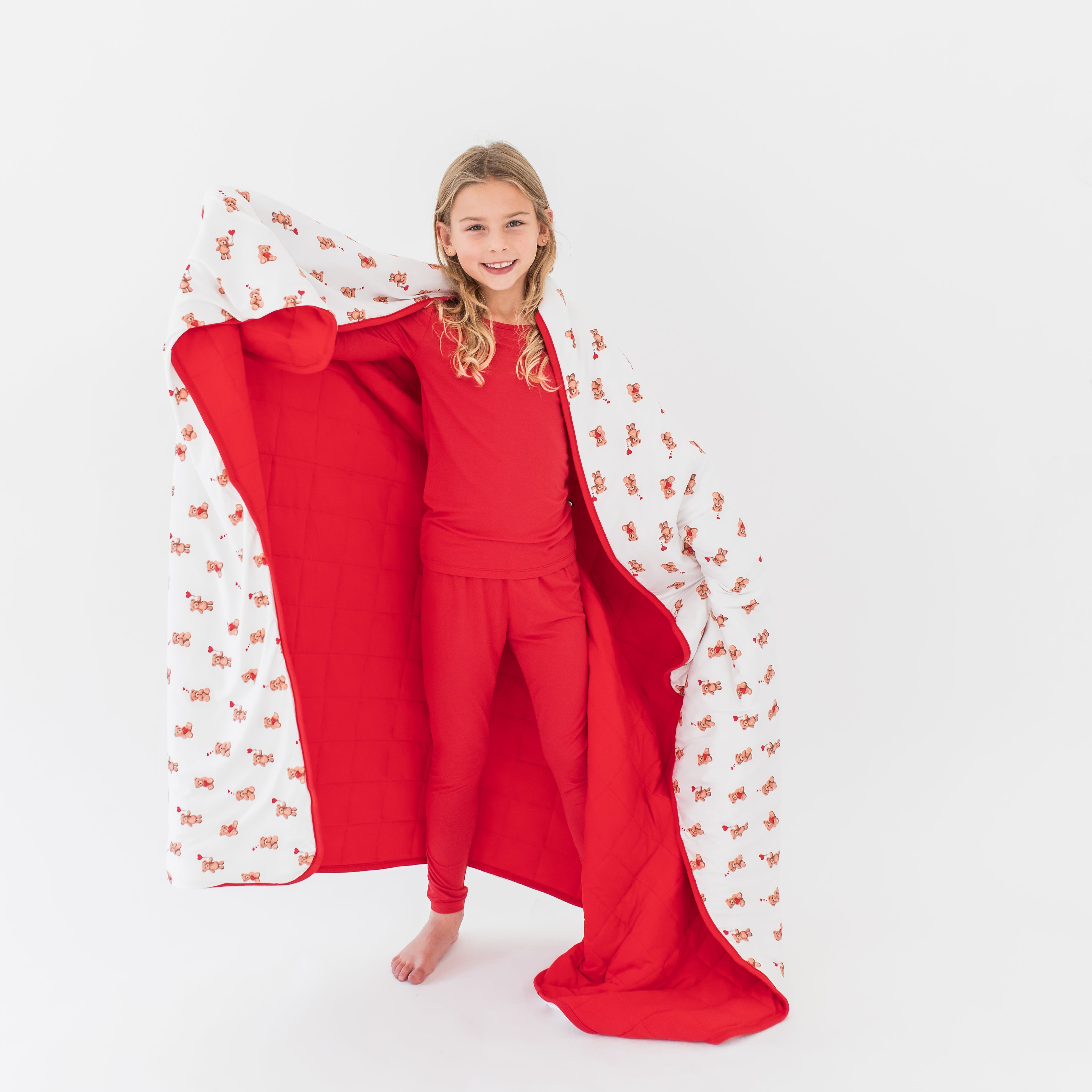 Young girl standing in front of a white background wearing cardinal pajamas with the Youth Blanket in Bear Hearts 2.5 around her shoulders showing the blankets red blanket and printed front