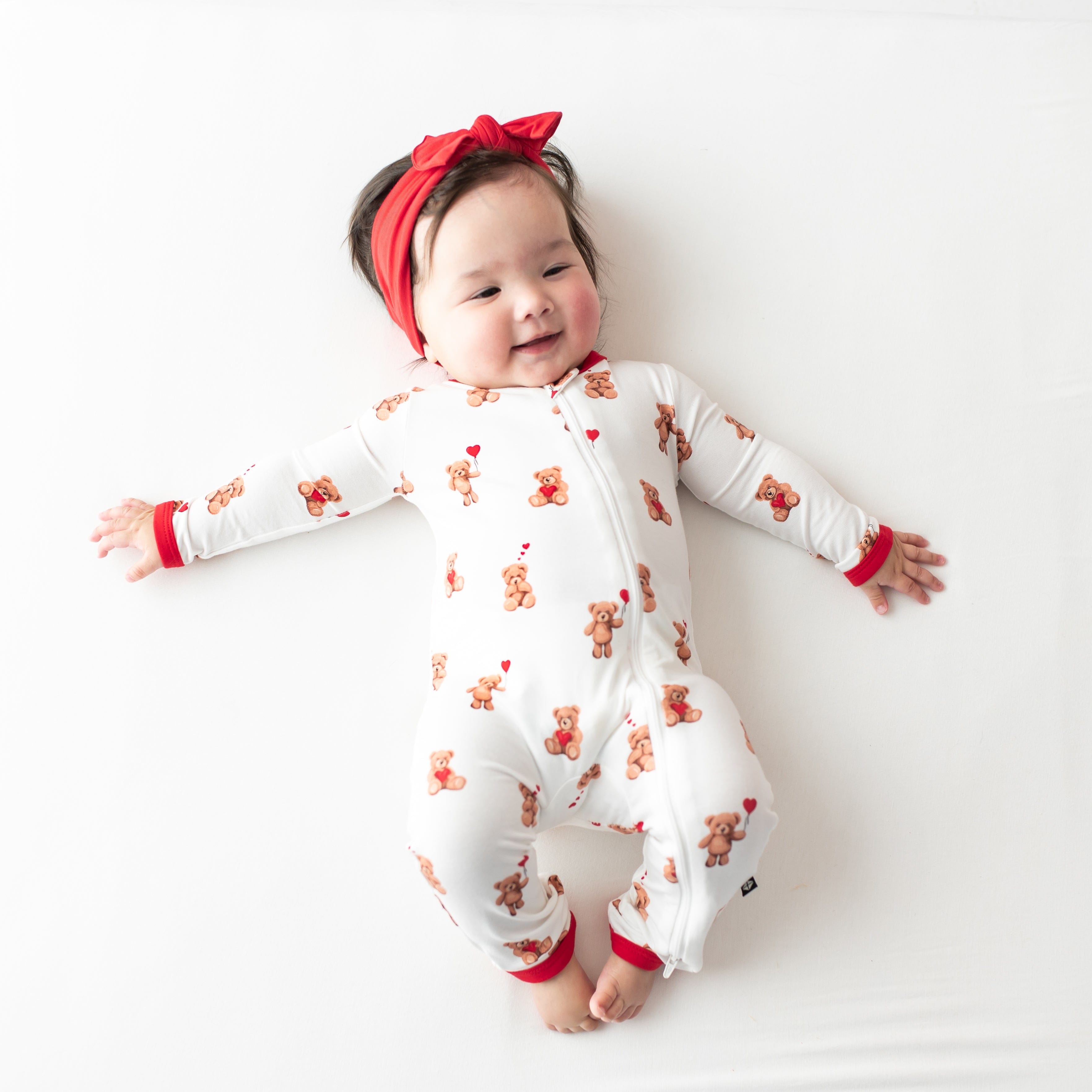Smiling infant lying down wearing the soft and breathable Zippered Romper in Bear Hearts paired with a Cardinal knotted bow headband