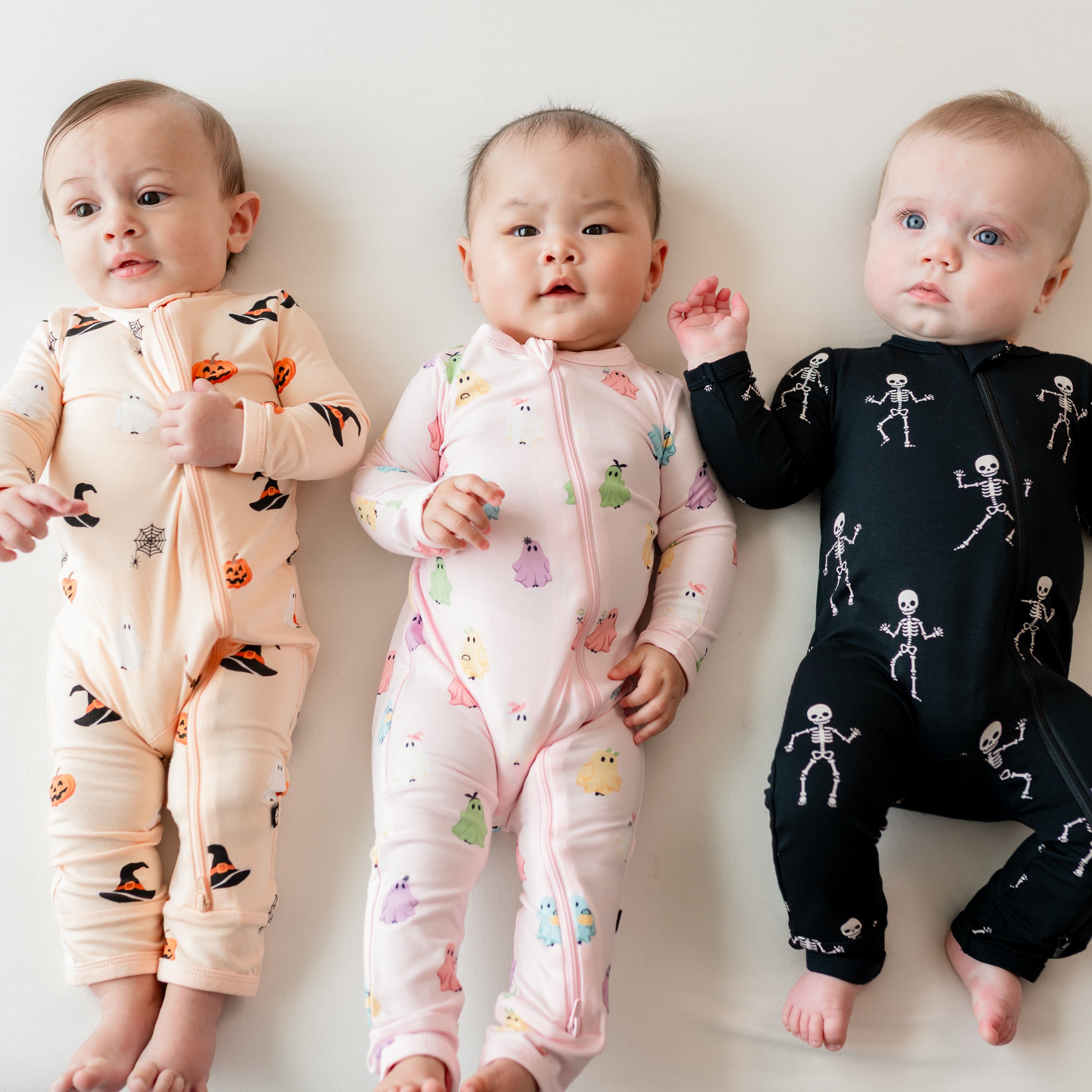 Three infant models laying side by side on a light colored blanket wearing the Zippered Romper in Pastel Ghost, Skeleton and Bewitched