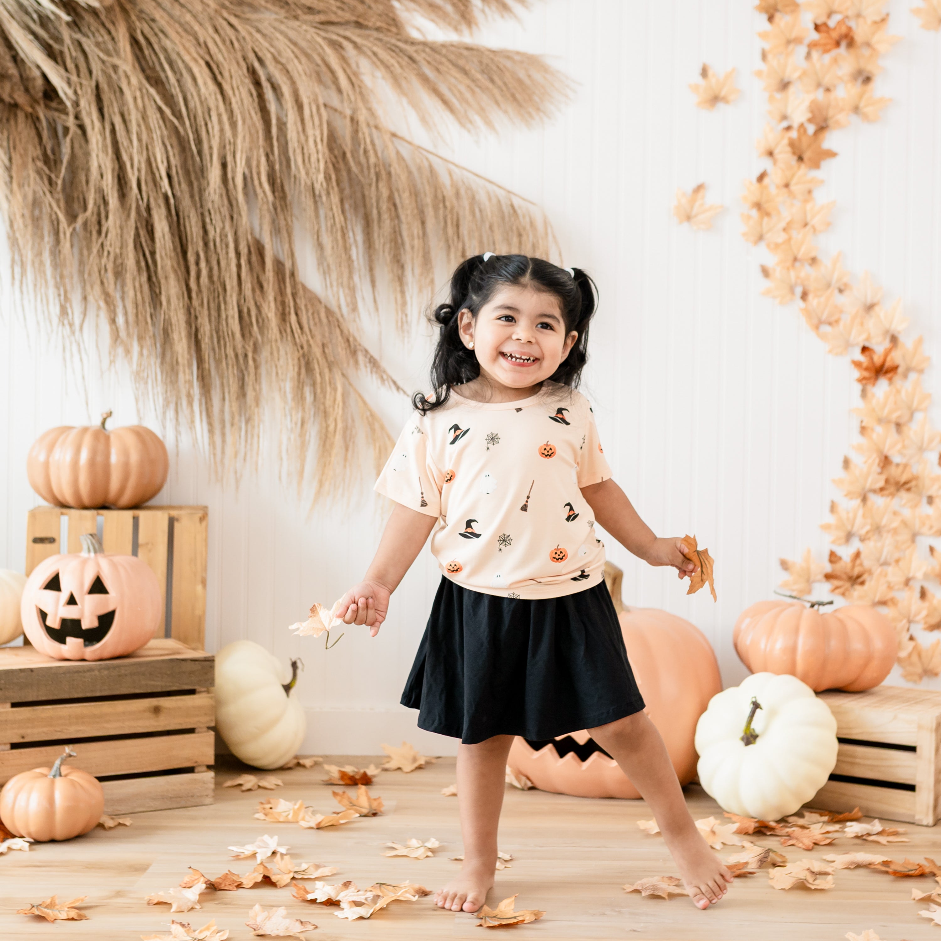 Toddler girl wearing the Toddler Basic Tee in Bewitched paired with a Midnight skort holding leaves in front of a fall inspired back drop