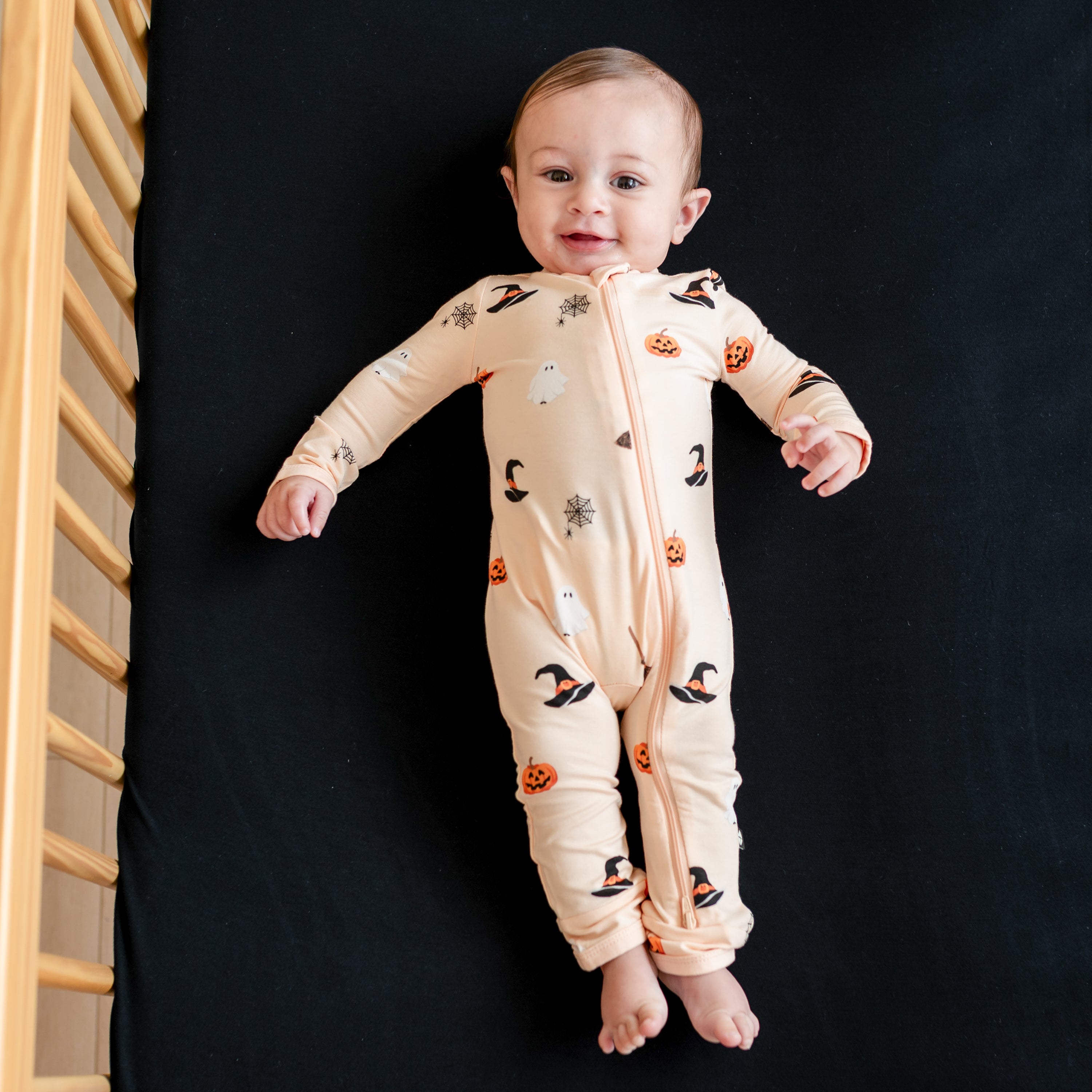 Infant laying in a crib on a Midnight crib sheet wearing the Zippered Romper in Bewitched