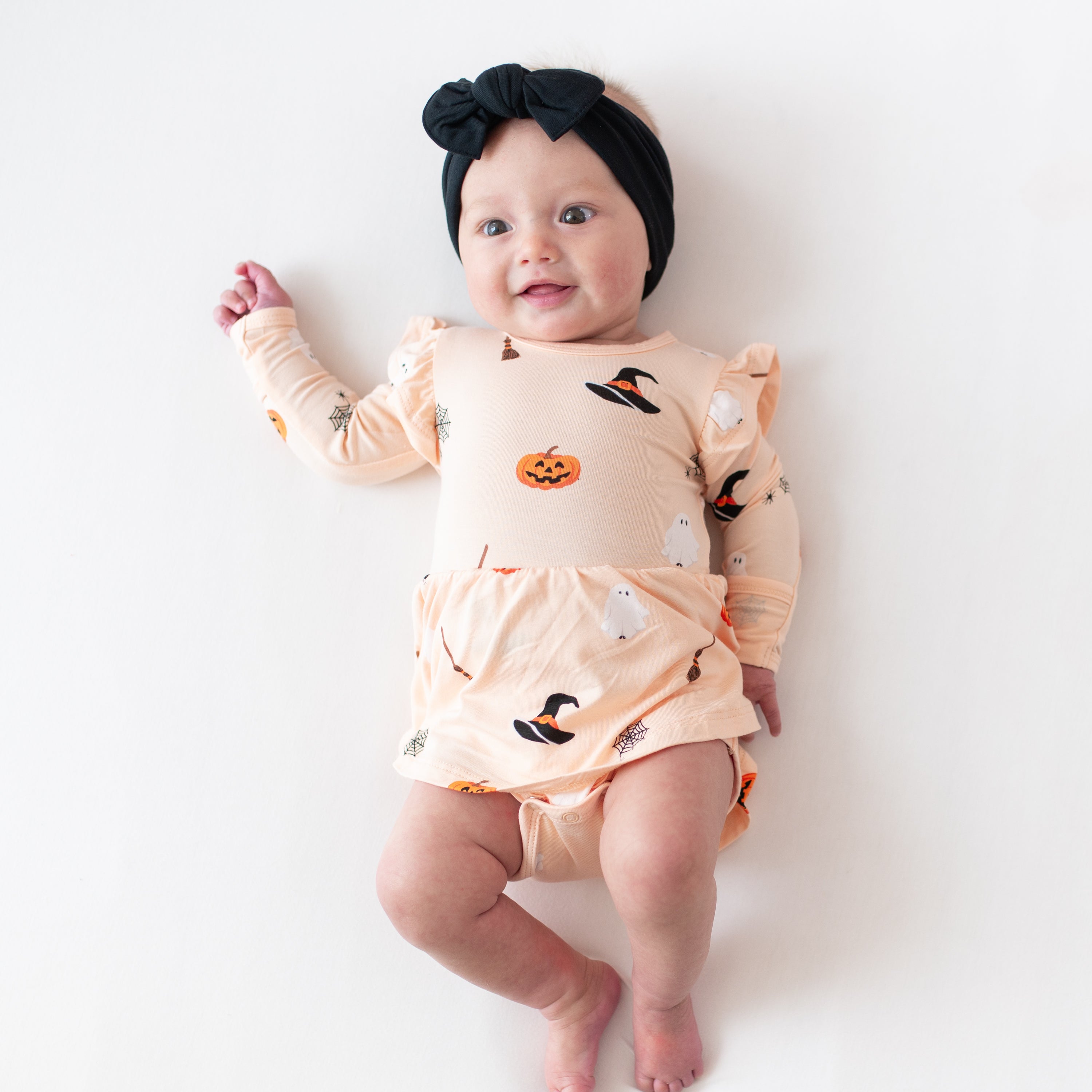 Baby wearing a Halloween-themed peach colored long sleeve twirl bodysuit dress and black bow with a white background