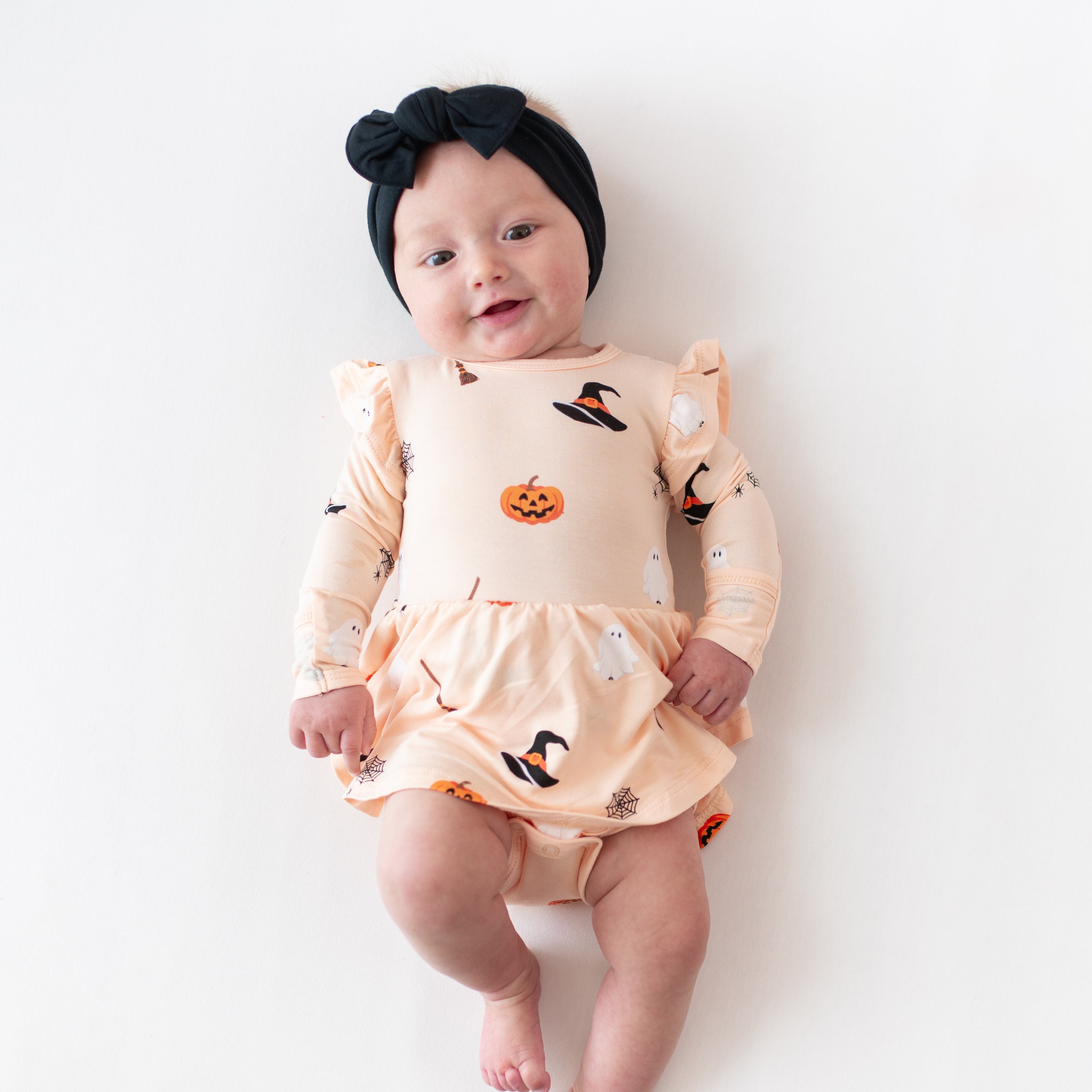 Baby wearing a Halloween-themed peach colored long sleeve bodysuit dress with pumpkins and ghosts on a white background