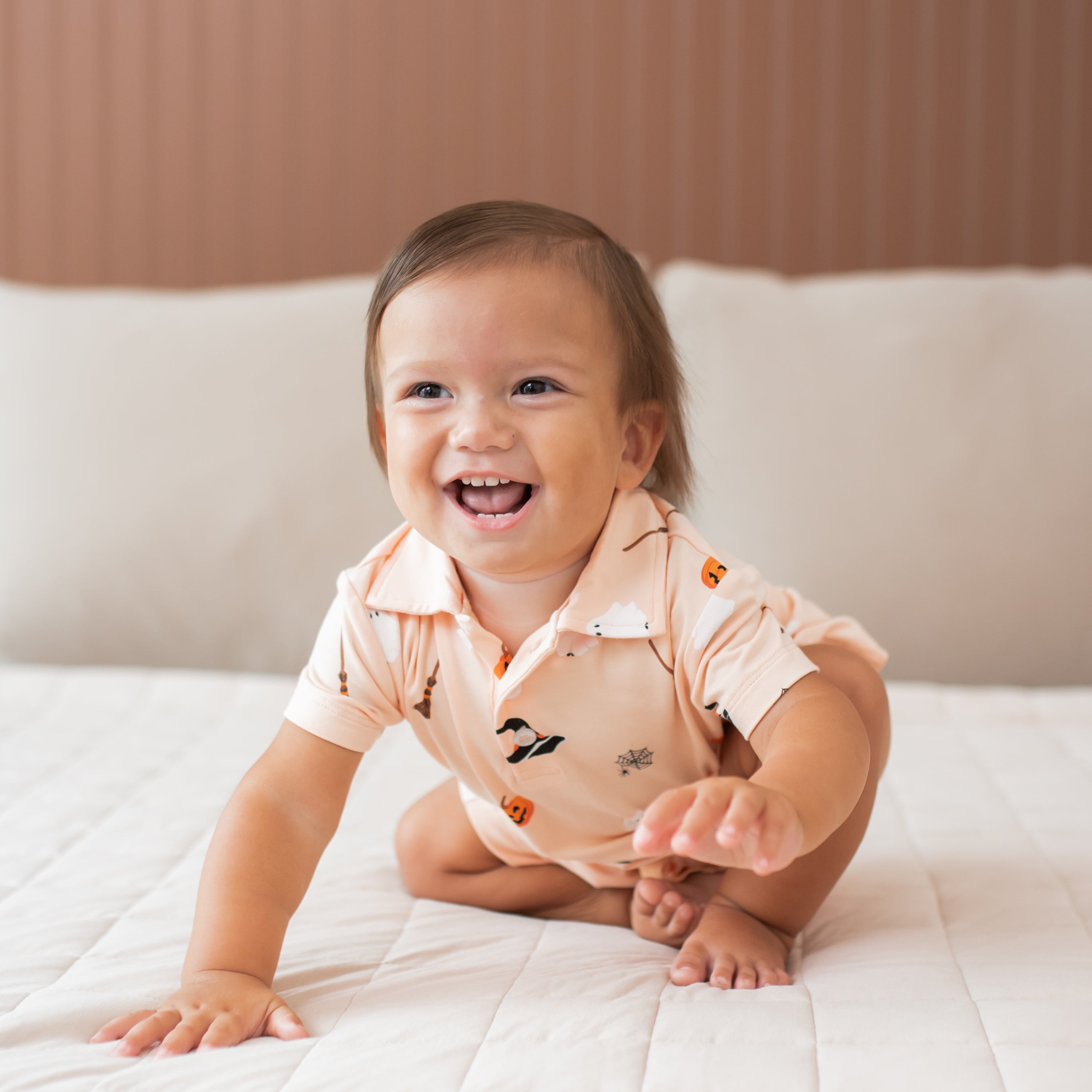 Baby sitting on a bed wearing a light orange colored polo shirt with halloween pattern