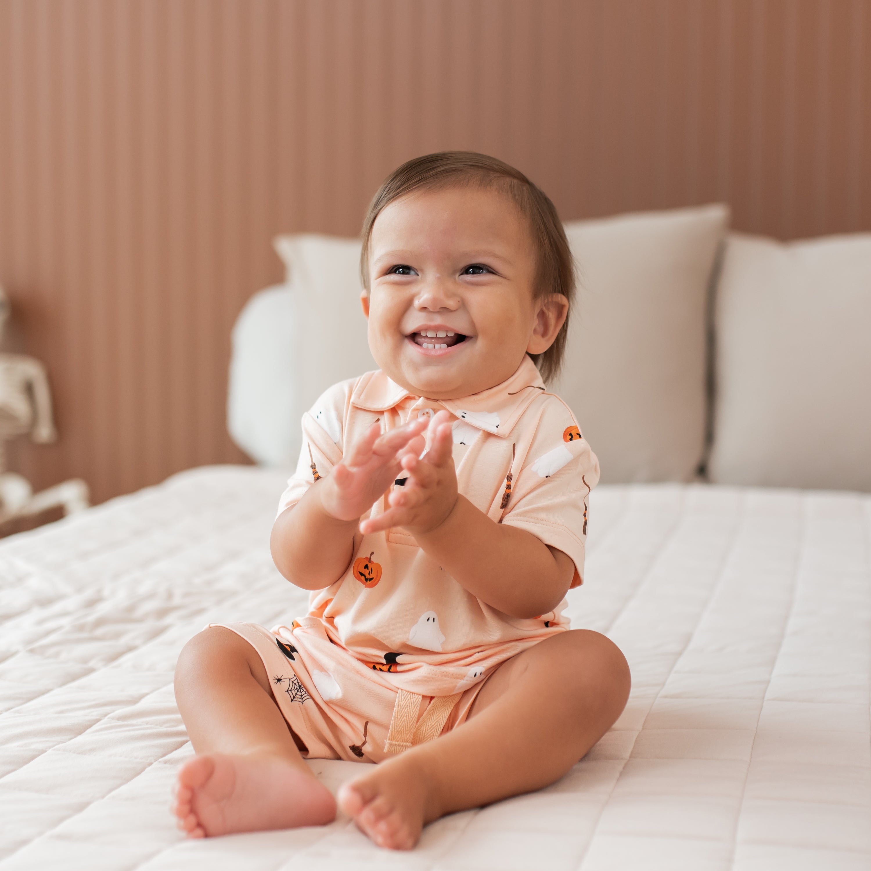 Baby in a halloween outfit sitting on a bed clapping hands with a smile