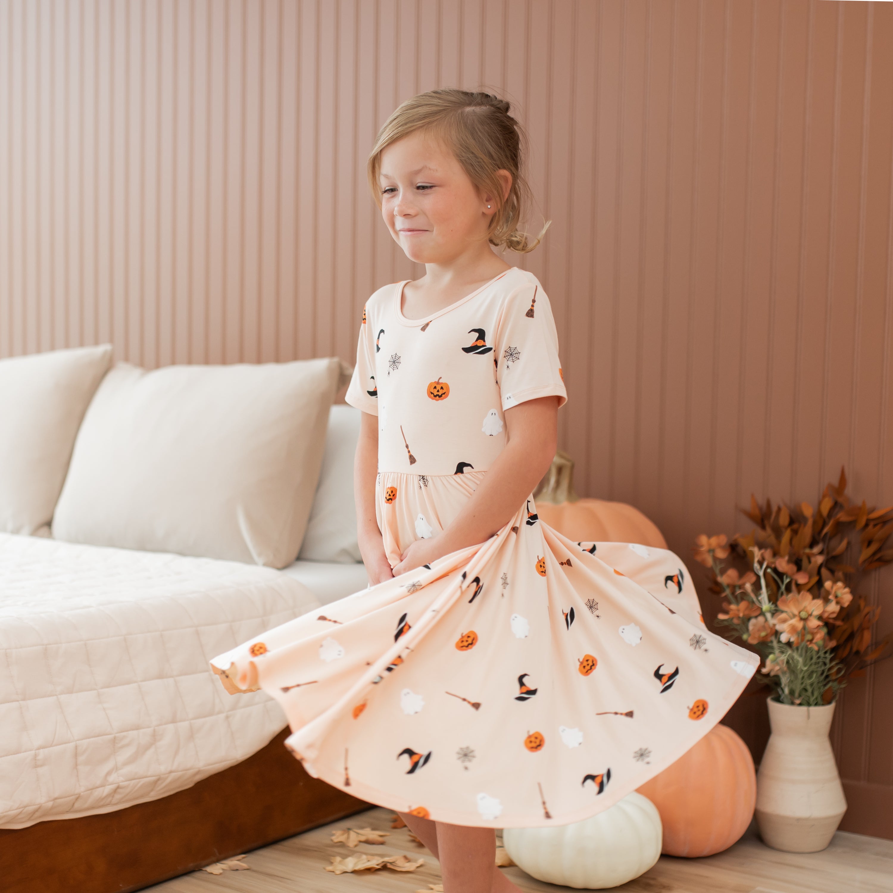 Young girl in a halloween patterned dress standing in a room with pumpkins and decorative items.
