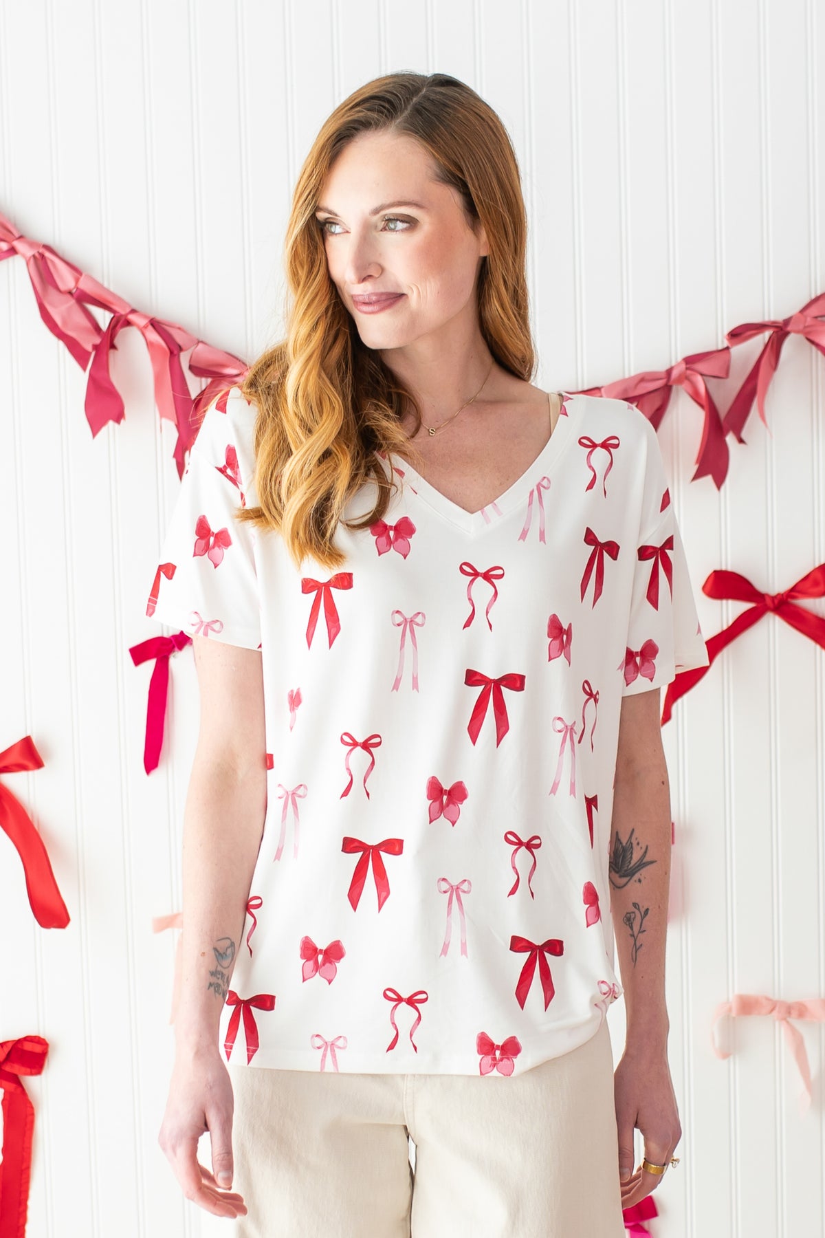 Female modeling the Women's Relaxed Fit V-Neck in Big Love Bow with light neutral pants standing in front of a bow garland hung on a white paneled wall