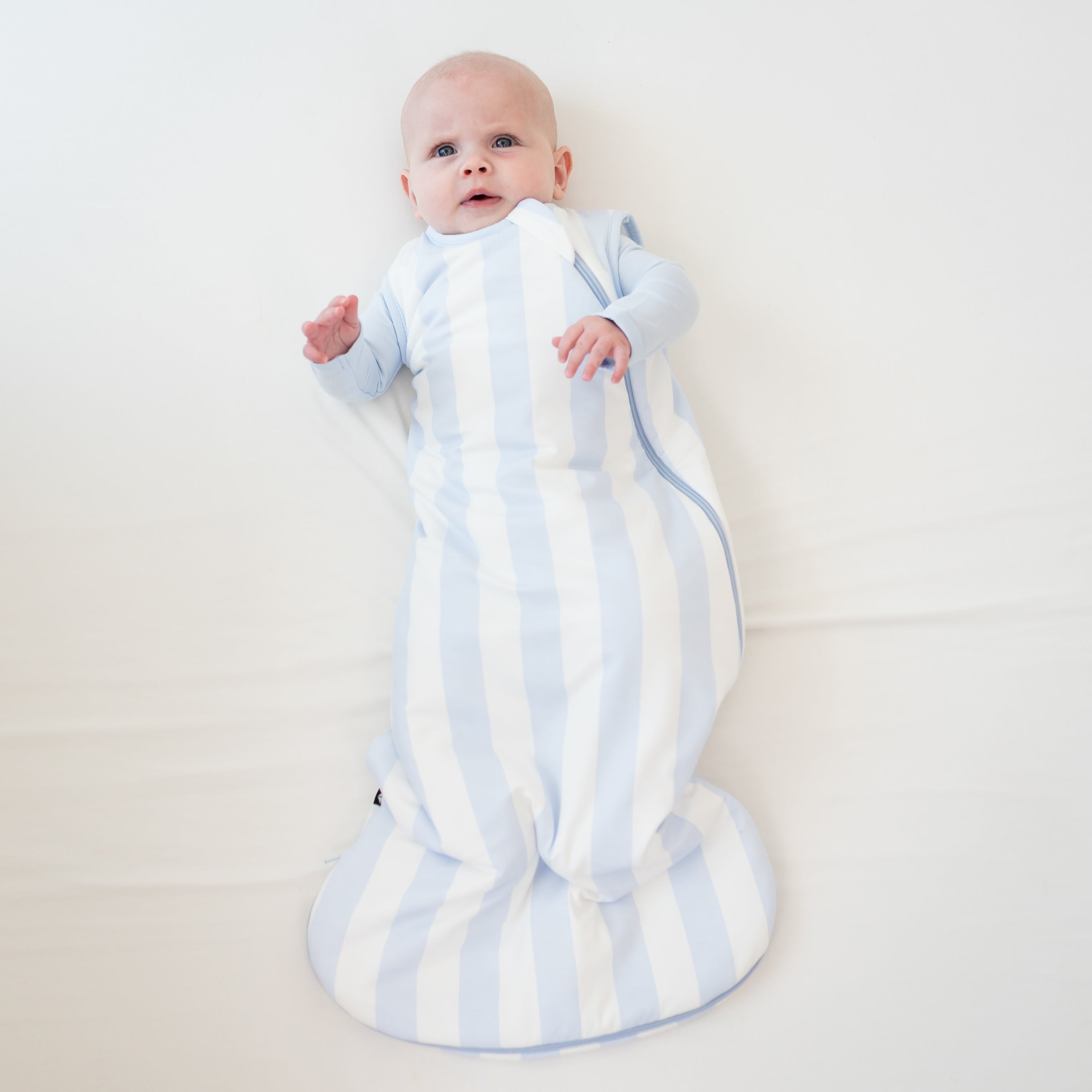Infant laying down wearing the Sleep Bag in Big Mist Stripe 1.0 with a Mist Romper on underneath