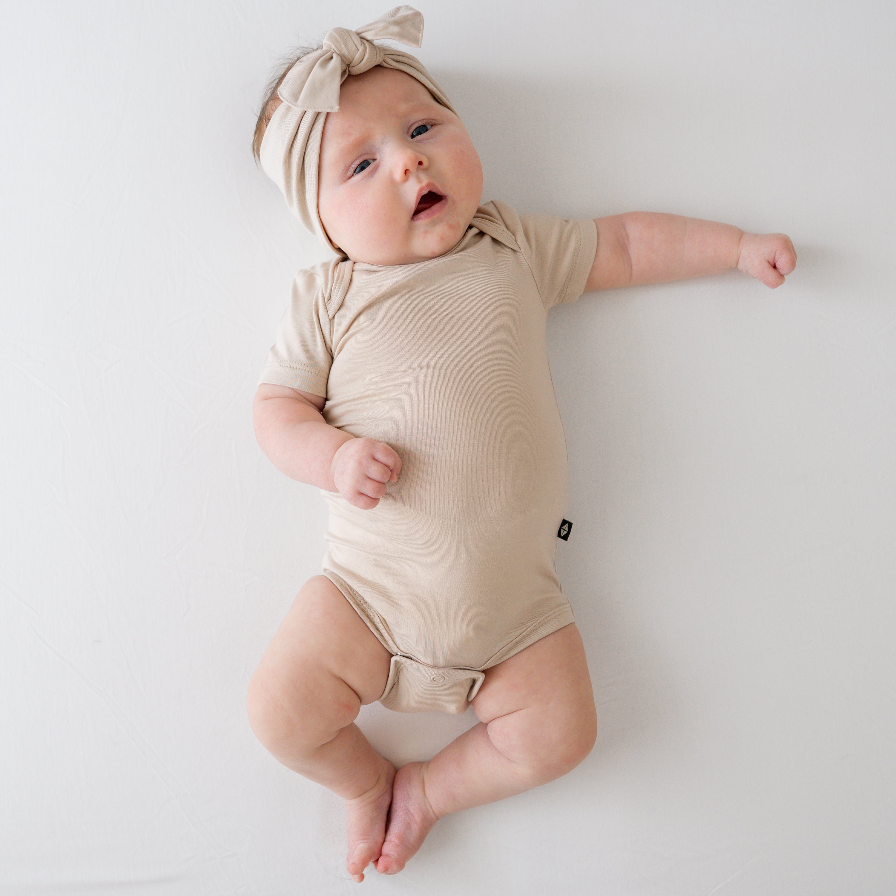 Baby in a Bisque onesie and matching bow lying on a white surface