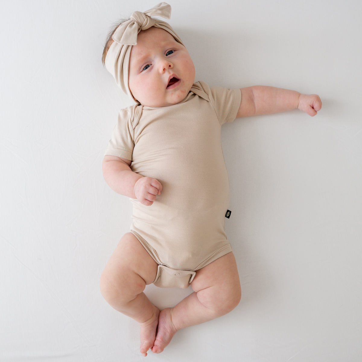 Baby in a Bisque onesie and matching bow lying on a white surface