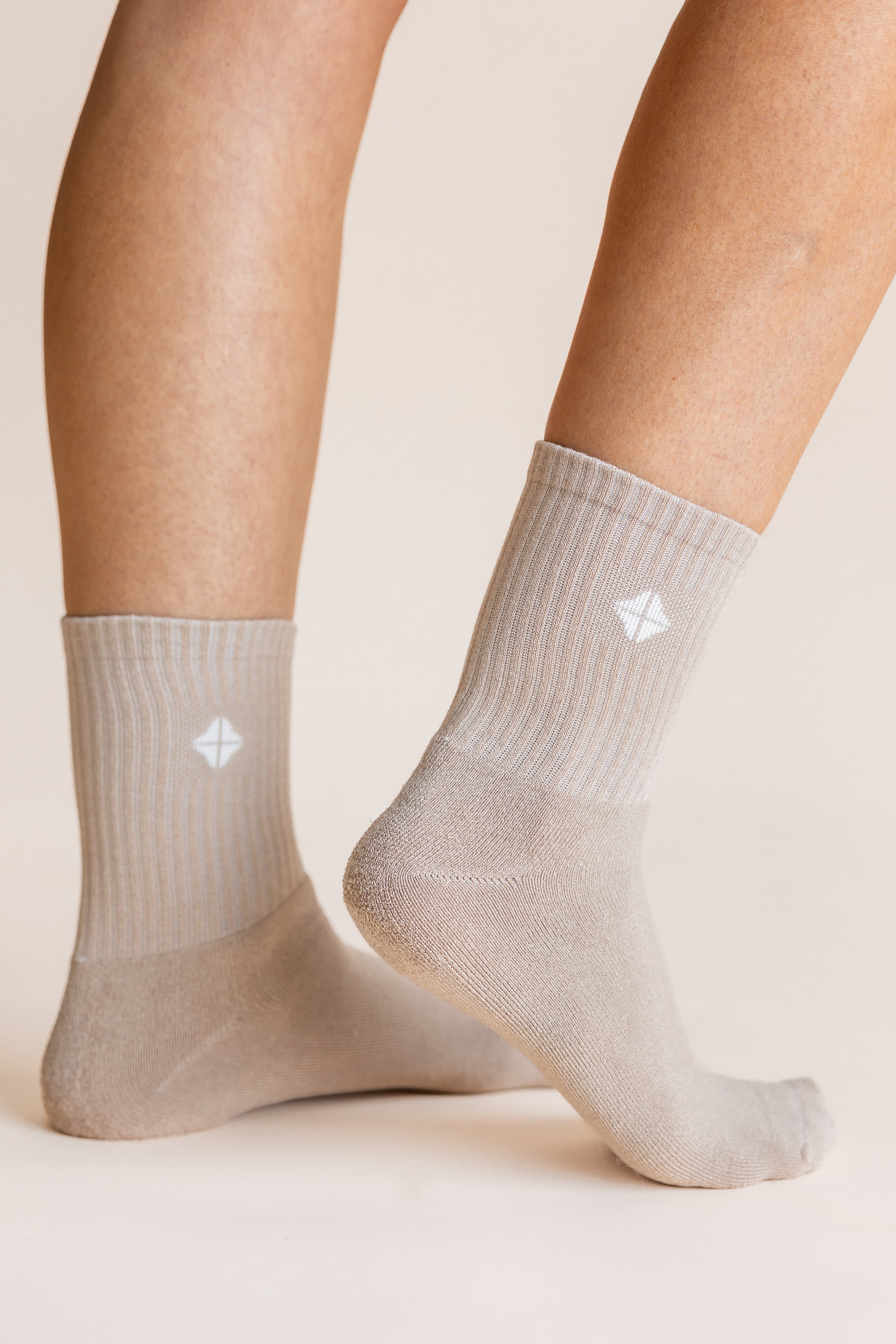 A close-up of a person's feet and lower legs wearing light beige crew socks with a small white geometric logo on the side, set against a solid warm beige background.