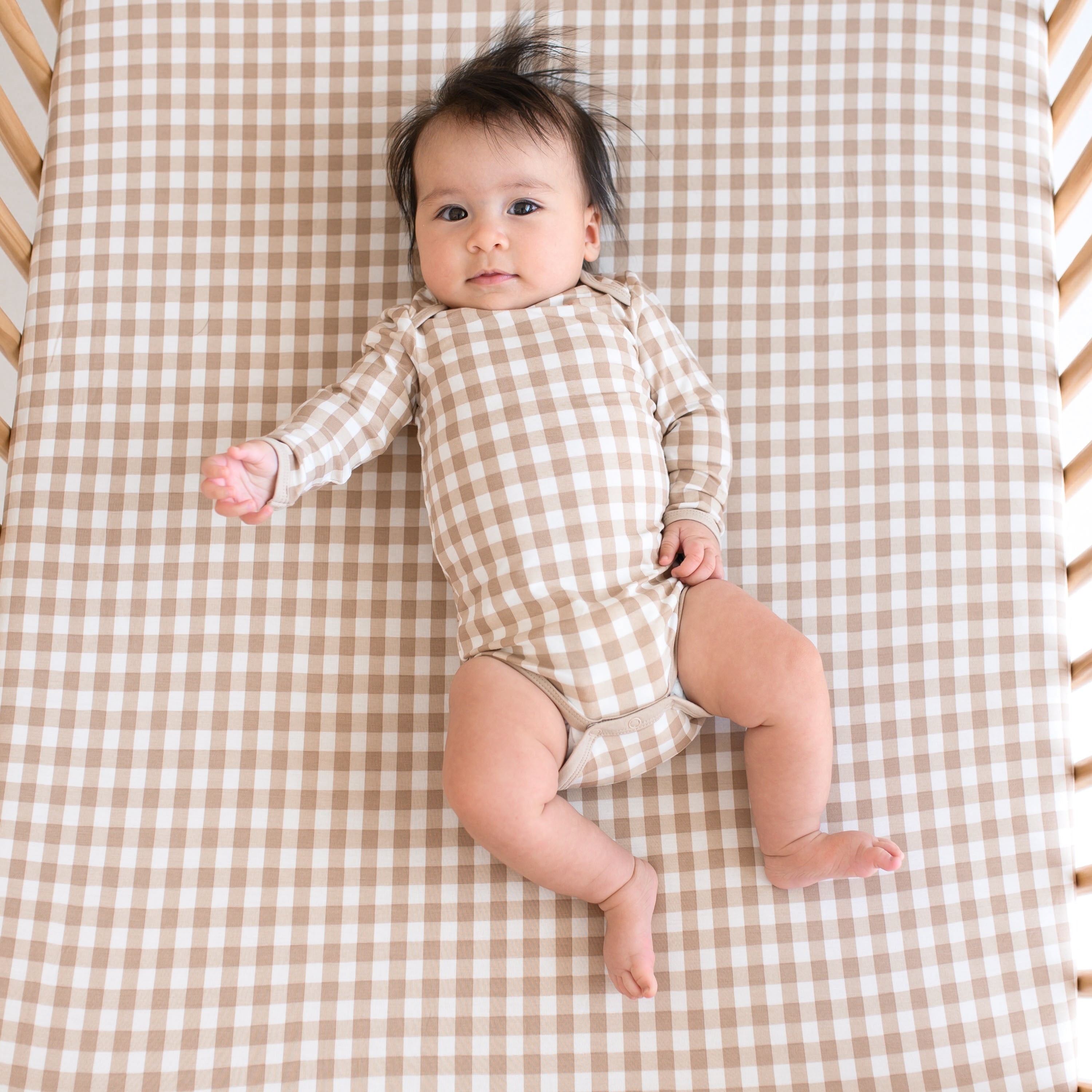 Infant laying in a crib on a Crib Sheet in Gingham Bisque wearing a matching long sleeve bodysuit