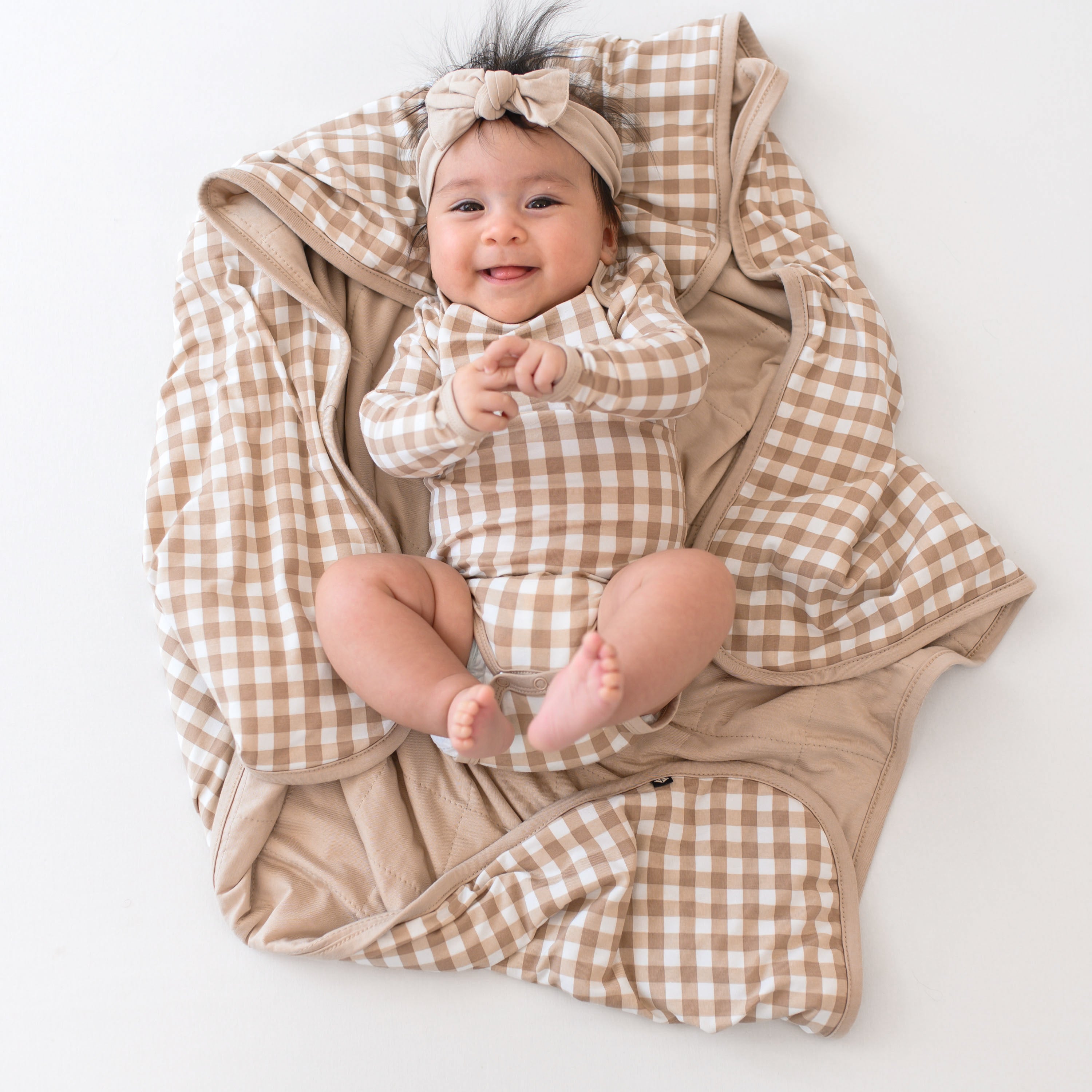 Infant laying on a Baby Blanket in Gingham Bisque wearing a matching long sleeve bodysuit and bisque knotted bow headband