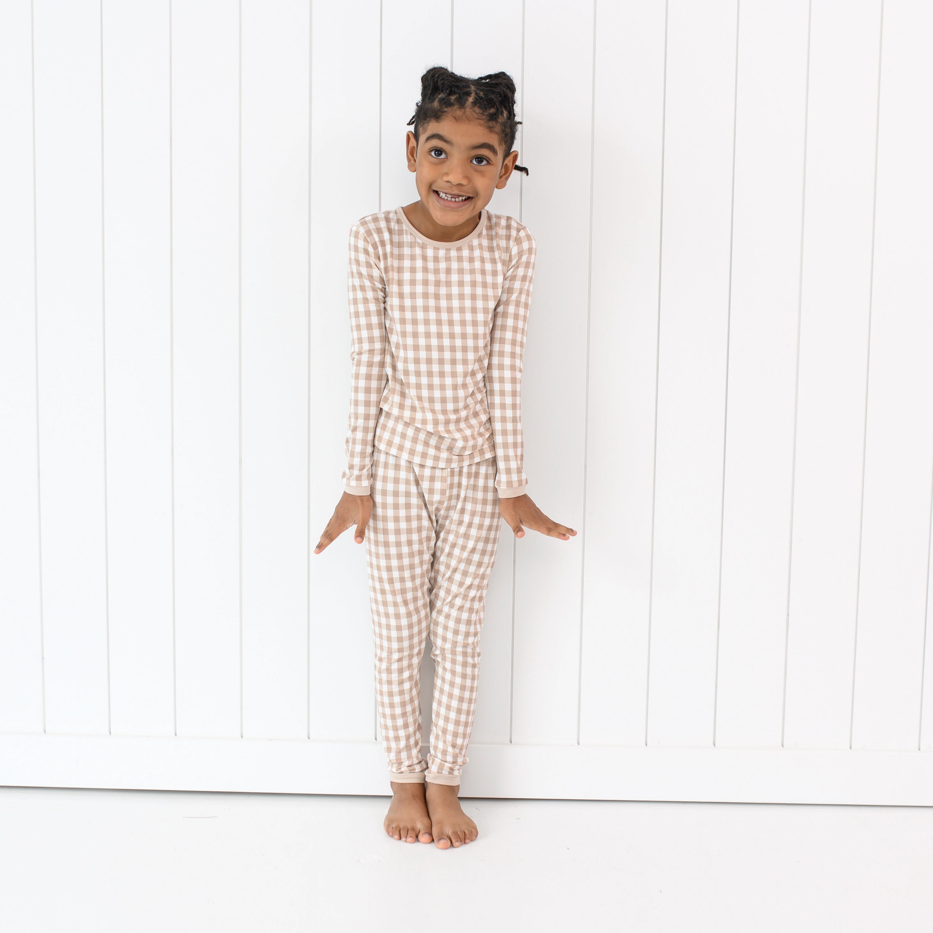 Young girl standing with our arms at her side wearing the Long Sleeve Pajamas in Gingham Bisque in front of a white paneled wall
