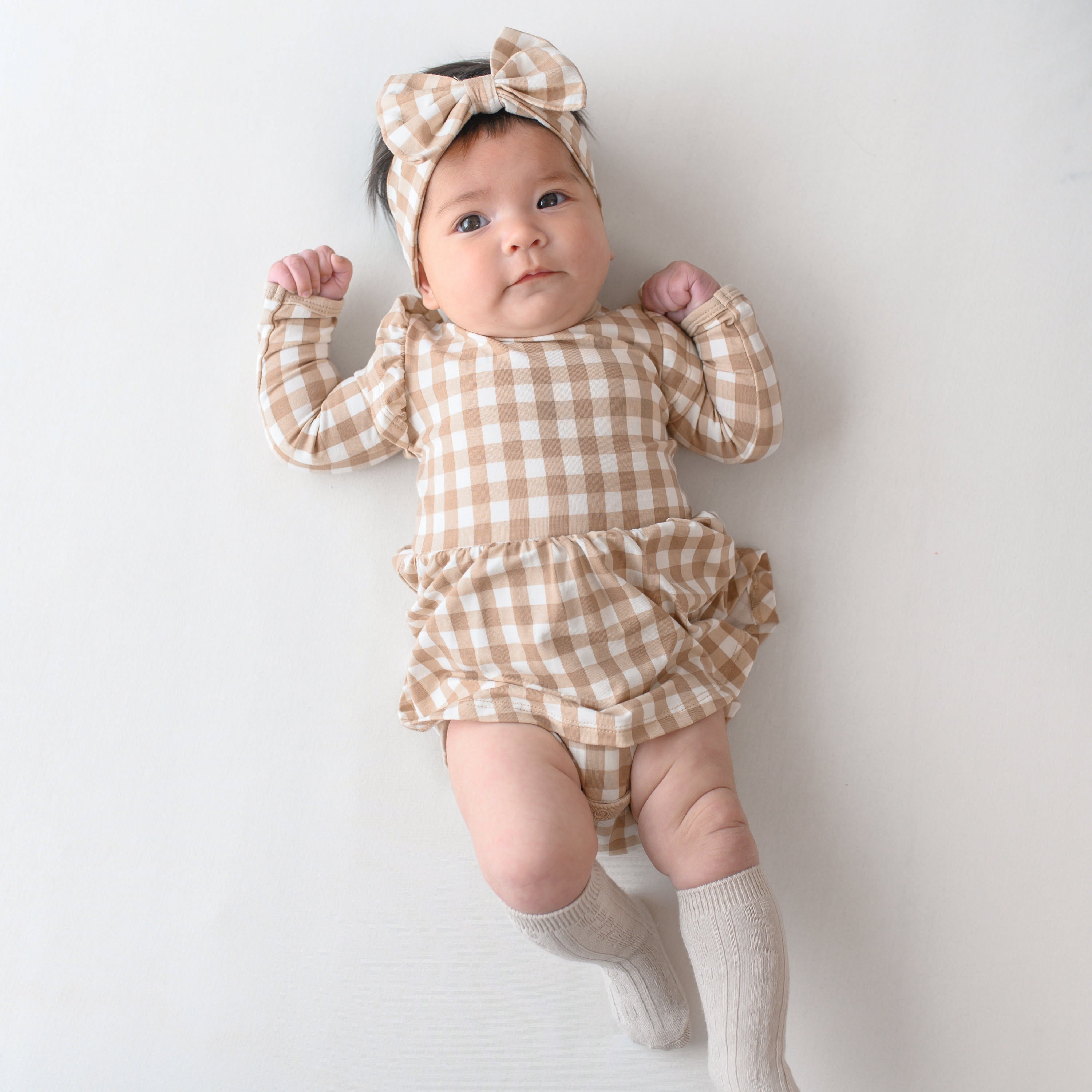 Infant girl laying on a light colored neutral background wearing the Long Sleeve Twirl Bodysuit Dress in Gingham Bisque paired with matching bow headband and Oat Knee high socks