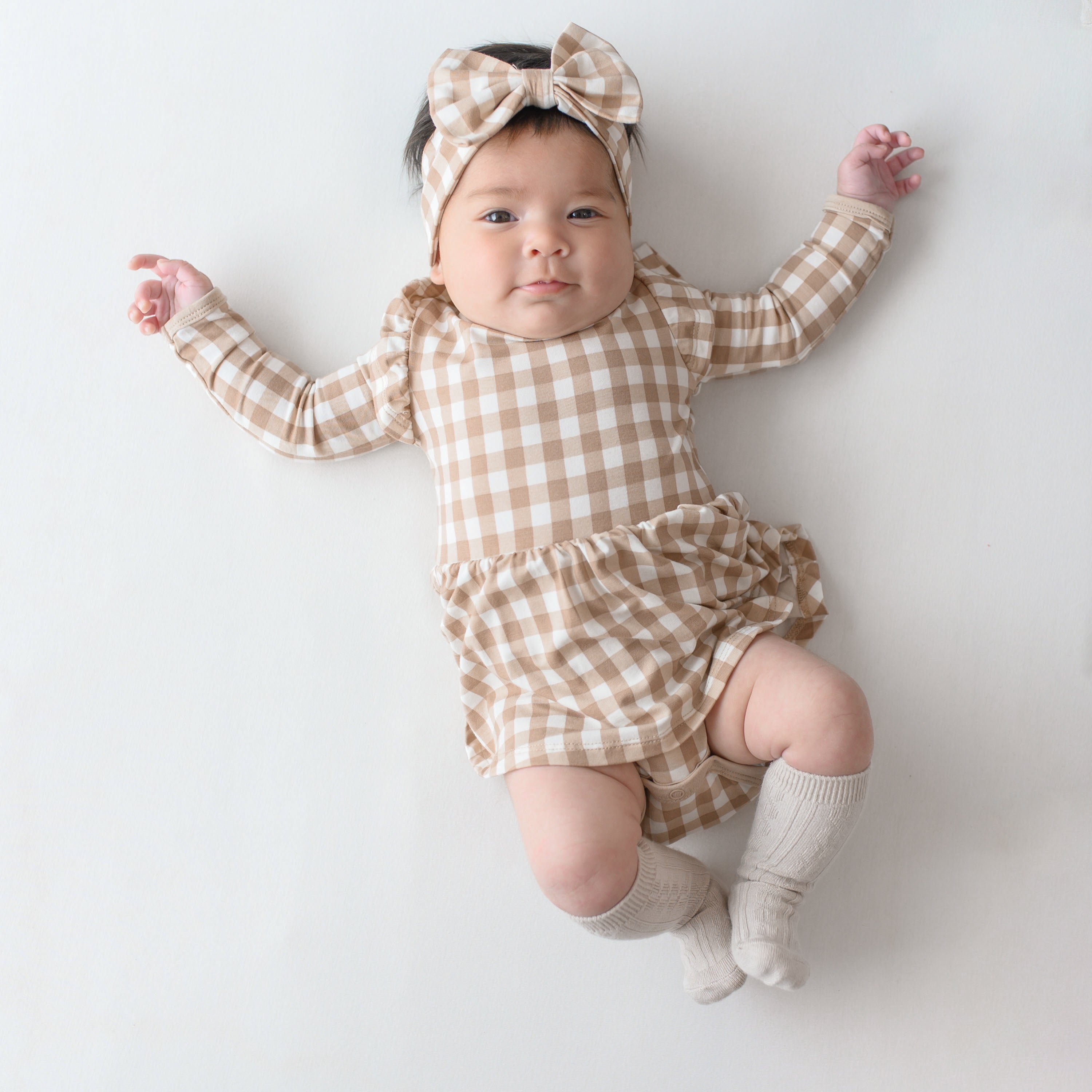 Smirking infant laying down on a light neutral colored backdrop wearing the Long Sleeve Twirl Bodysuit Dress in Gingham Bisque and matching bow headband with Oat Knee High socks