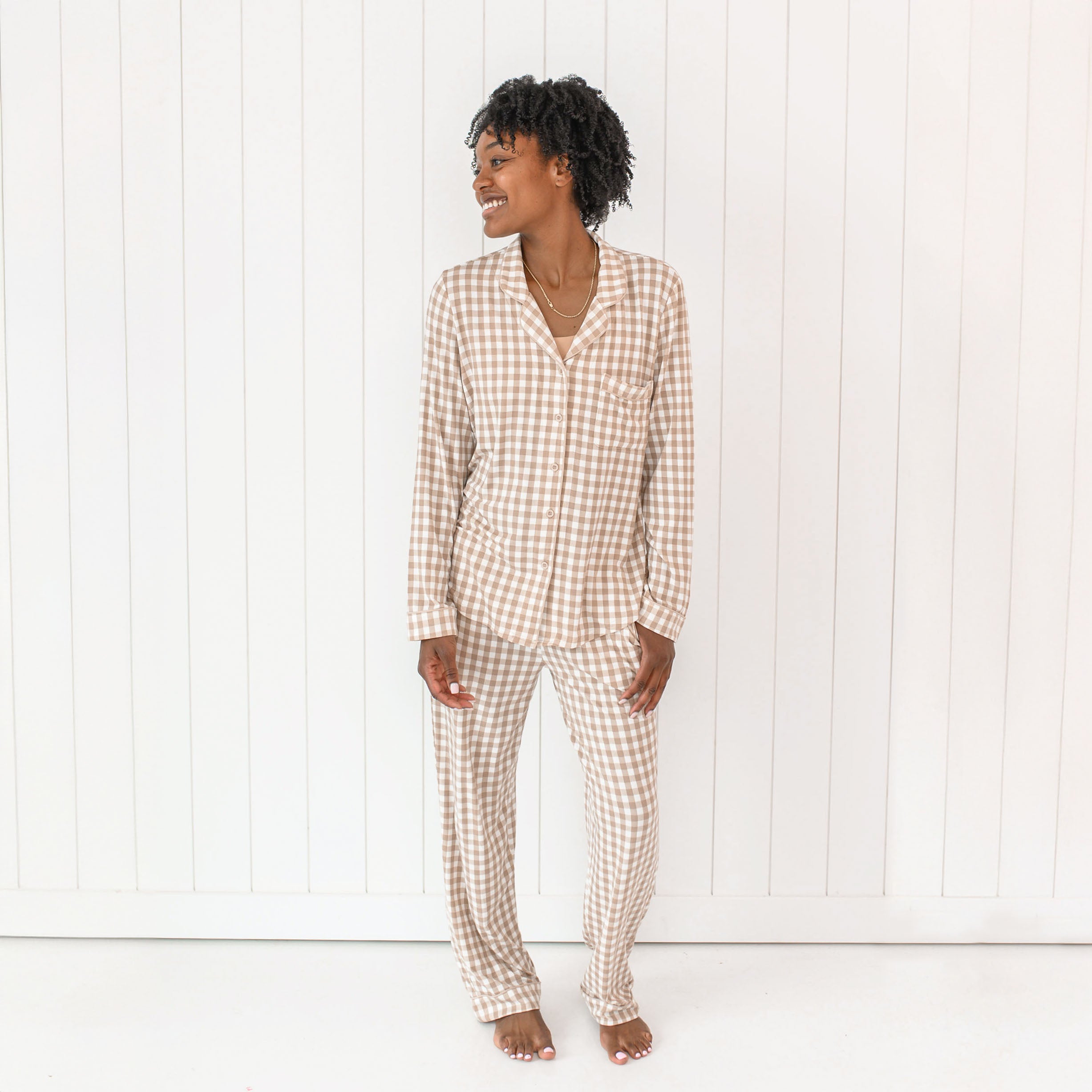 Female model wearing the Long-Sleeved Women's Pajama Set in Gingham Bisque standing in front of a white paneled wall with one hand in the pajama pant pocket
