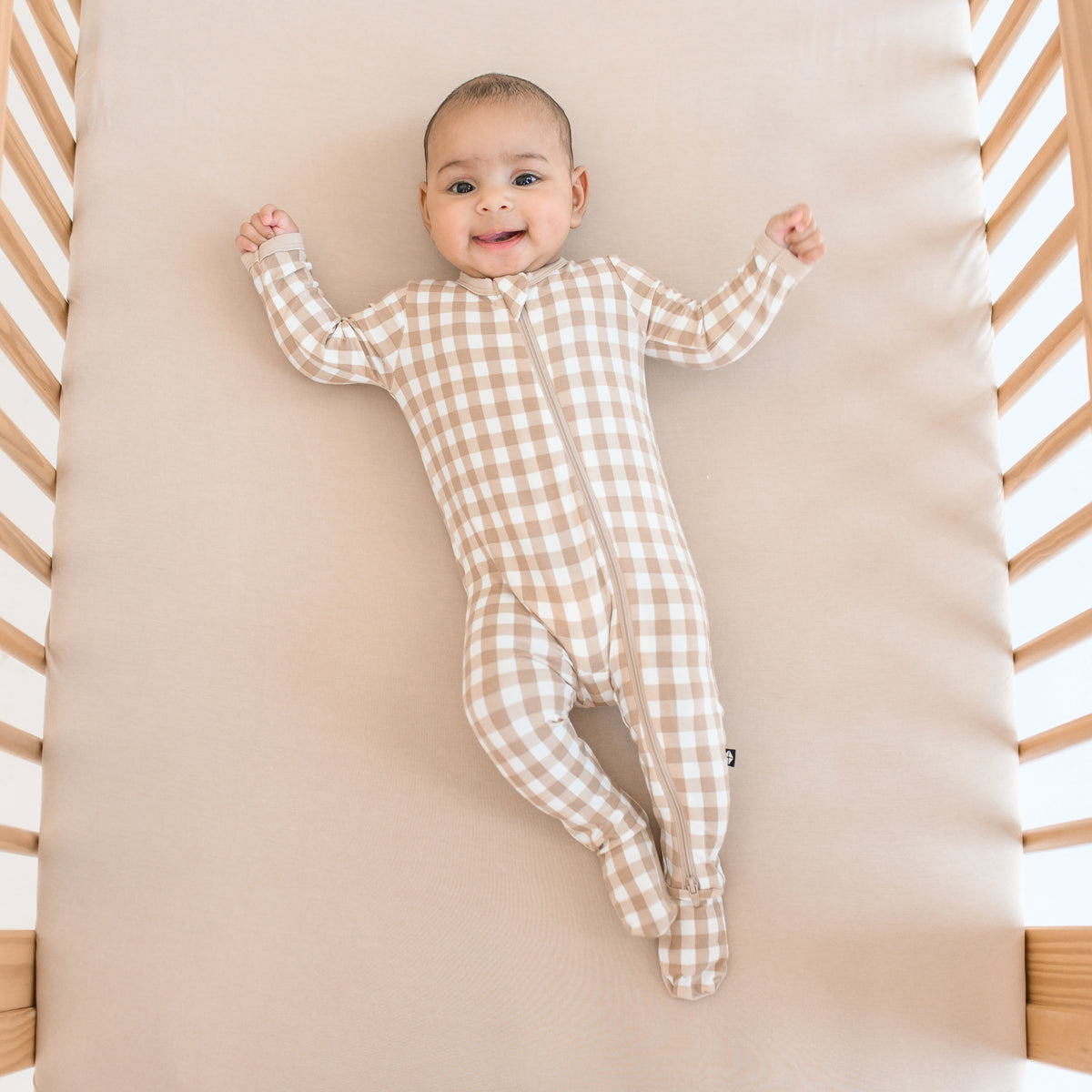 Infant laying in a crib on a Bisque crib sheet wearing the Zippered Footie in Gingham Bisque