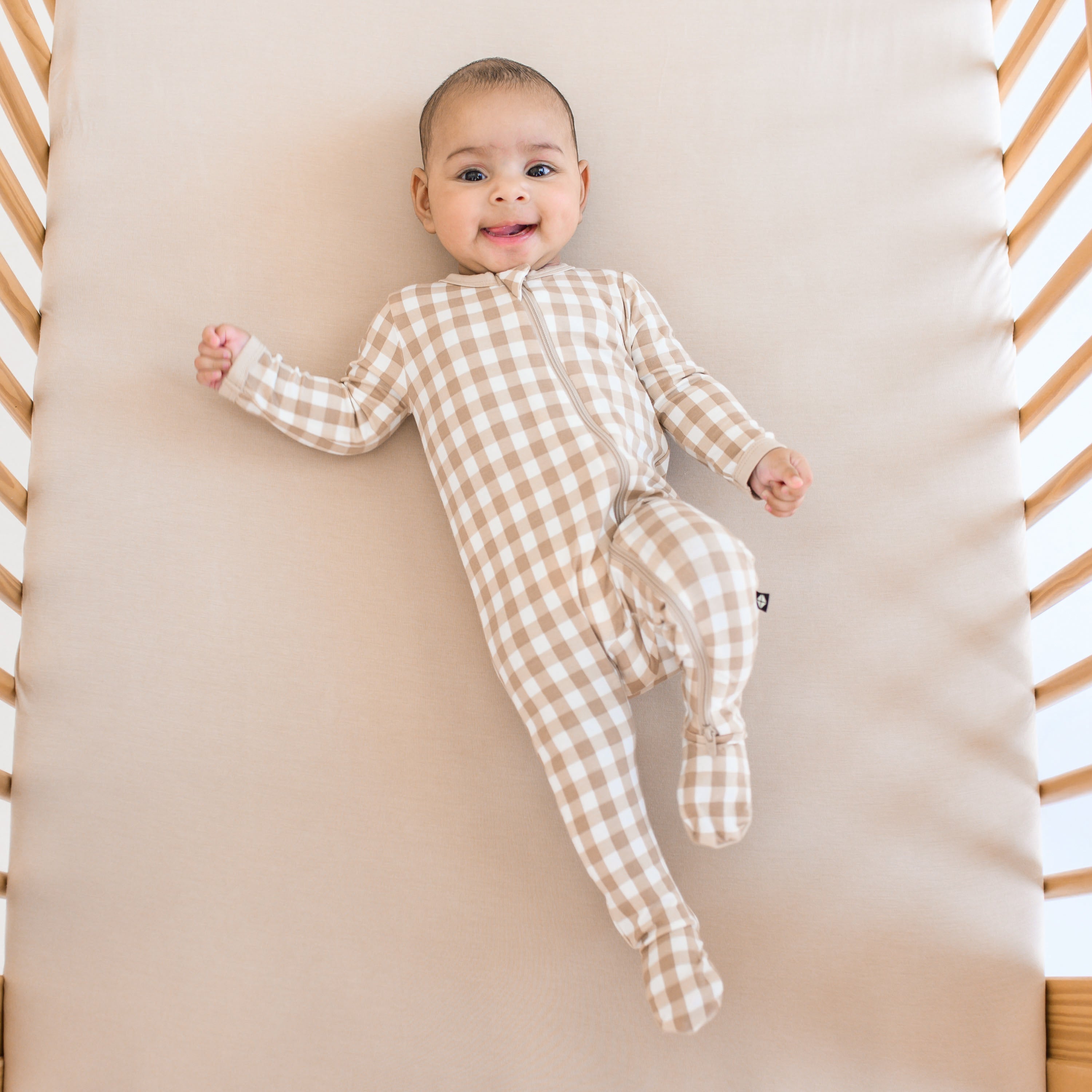 Infant laying in a crib laying on a Bisque crib sheet wearing the Zippered Footie in Gingham Bisque