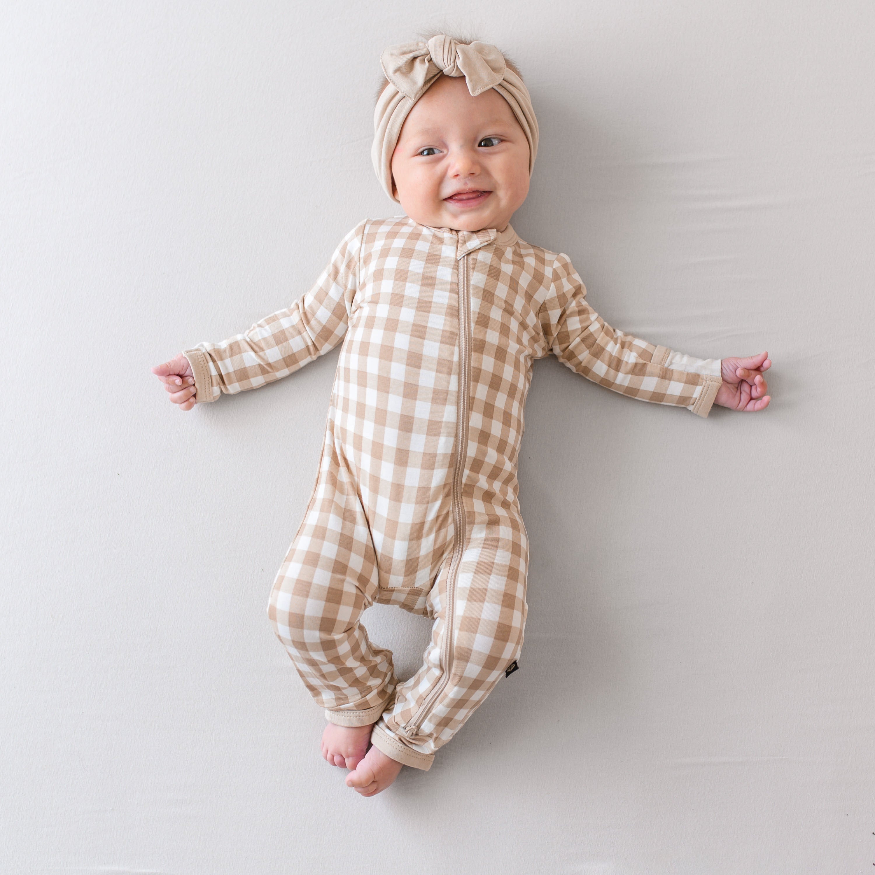 Infant laying down wearing the Zippered Romper in Gingham Bisque paired with a Bisque Knotted Bow Headband