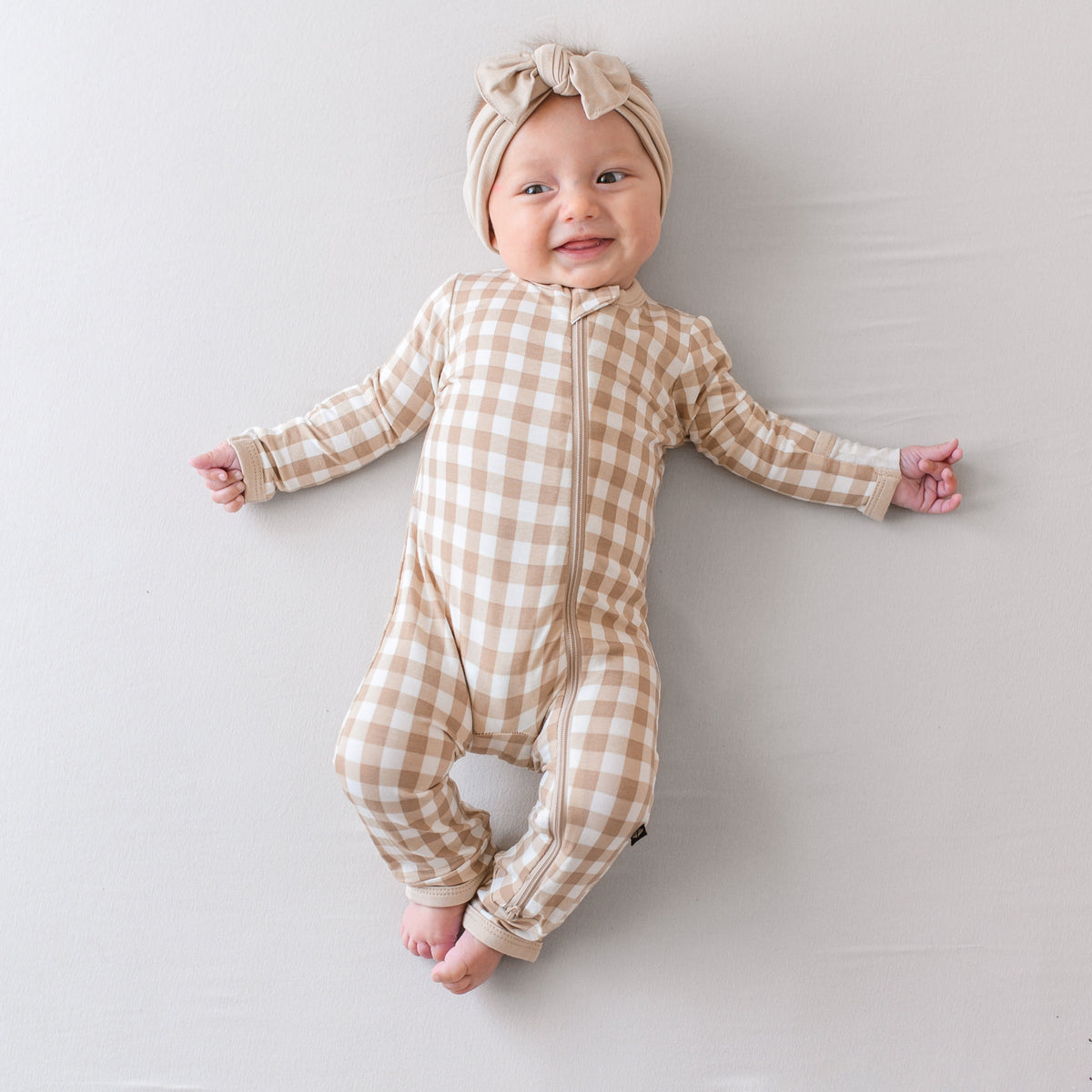 Infant laying down wearing the Zippered Romper in Gingham Bisque paired with a Bisque Knotted Bow Headband