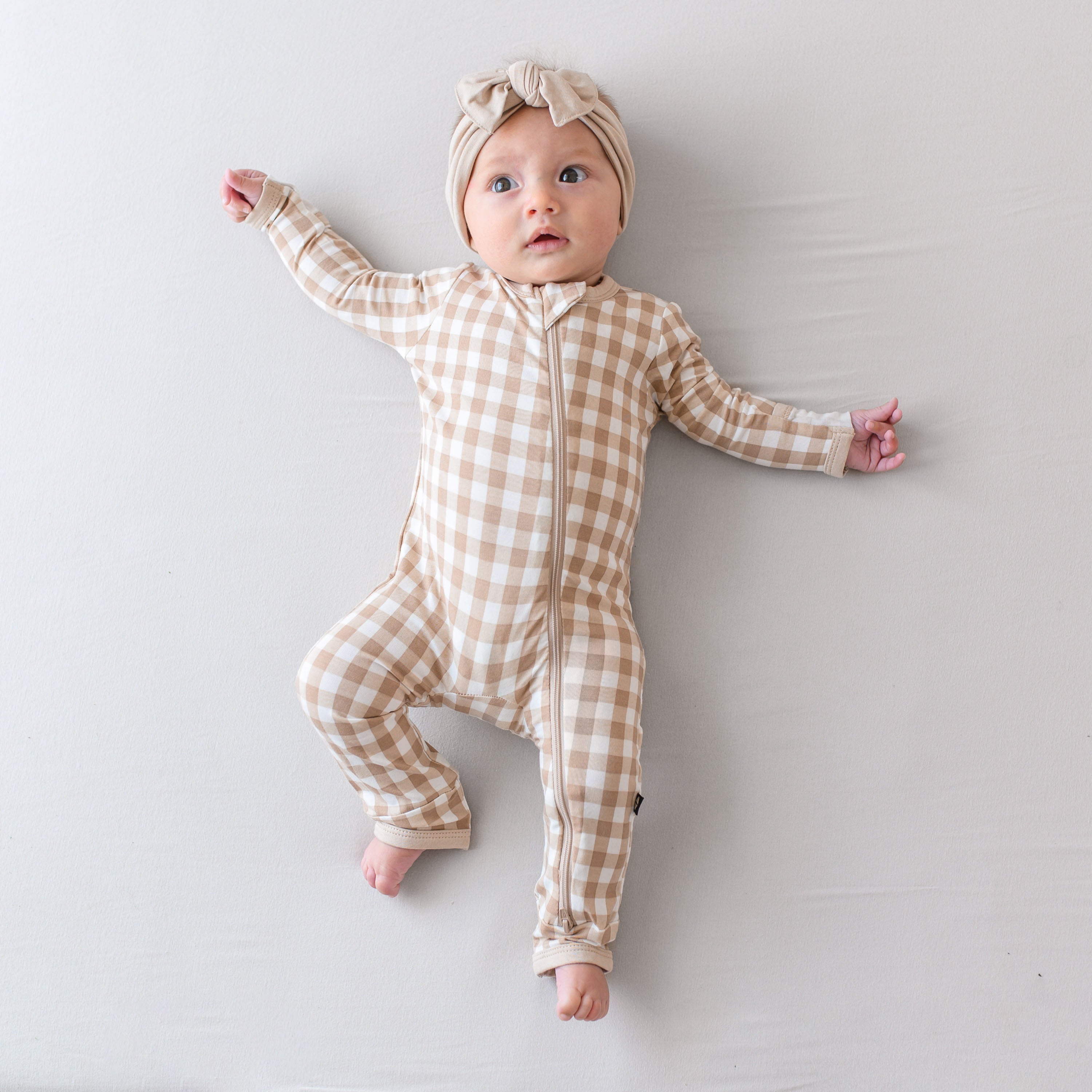 Infant laying down wearing the Zippered Romper in Gingham Bisque and Knotted Bow headband in Bisque