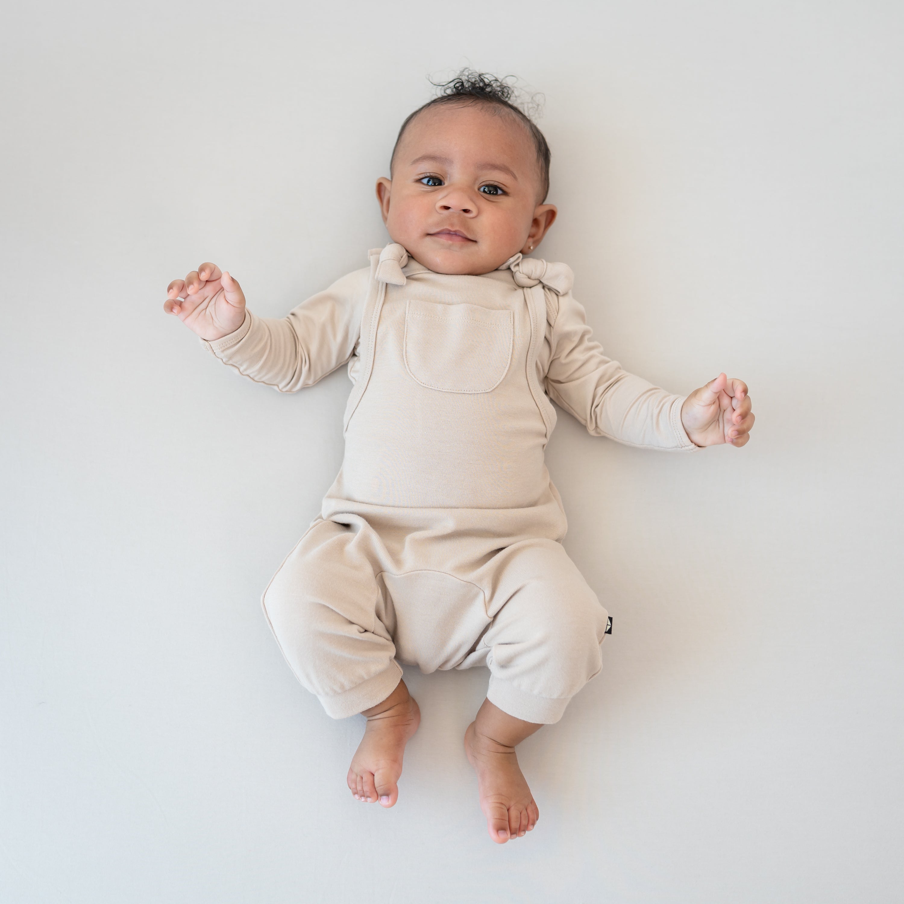 Infant laying down wearing the Bamboo Jersey Overall in Bisque with matching long sleeve bodysuit on underneath