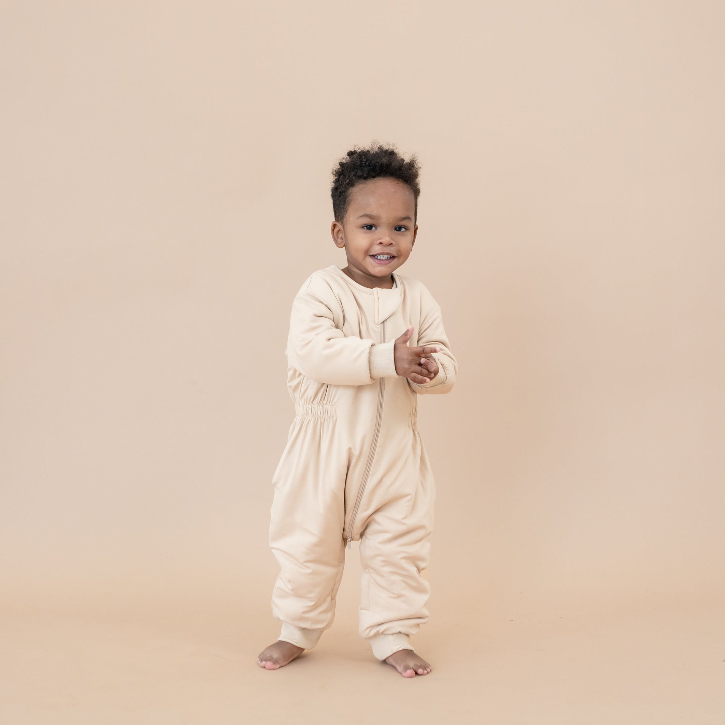 Young boy walking wearing the Cozy Playsuit in Bisque in front of a light taupe backdrop