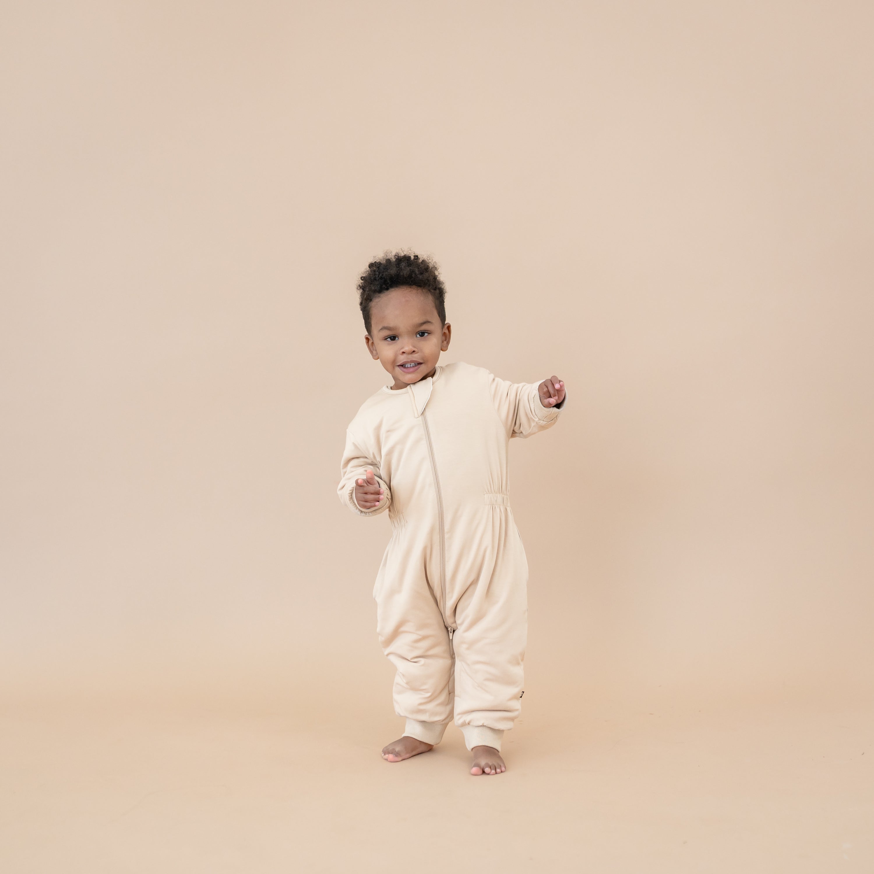 Young boy walking wearing the Cozy Playsuit in Bisque in front of a light taupe backdrop