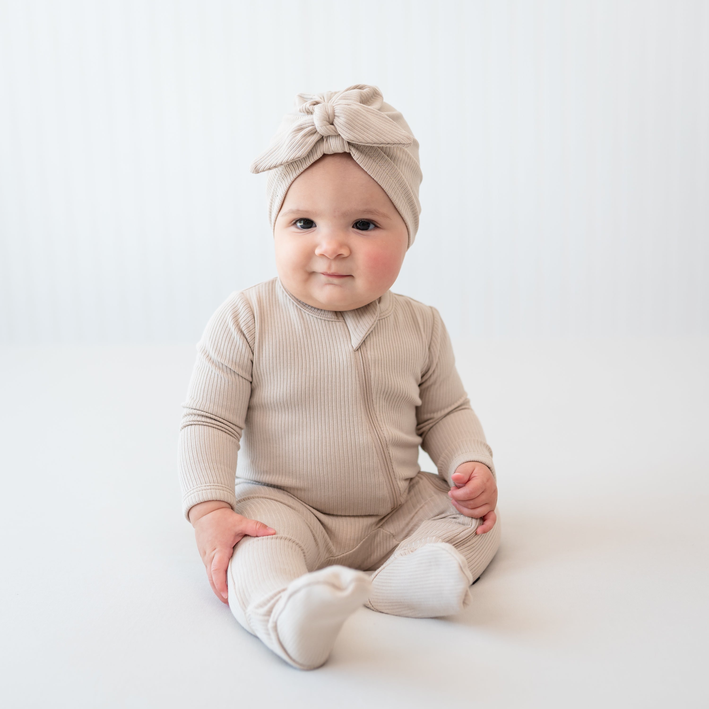 Infant girl sitting in front of a neutral backdrop wearing the Ribbed Zipper Footie in Bisque and matching ribbed headwrap