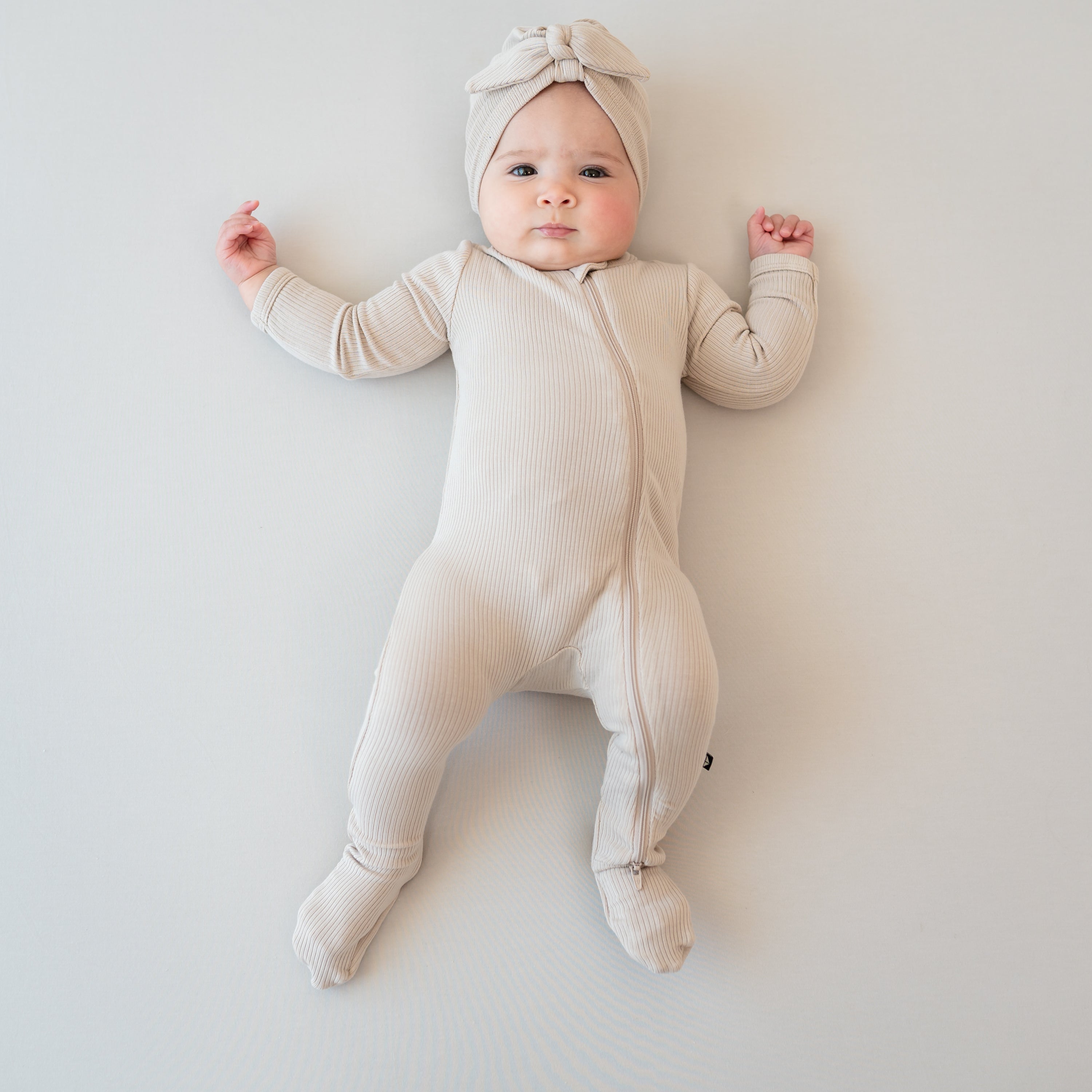 Infant girl laying down on a light neutral background wearing the Ribbed Zipper Footie in Bisque paired with a matching ribbed headwrap