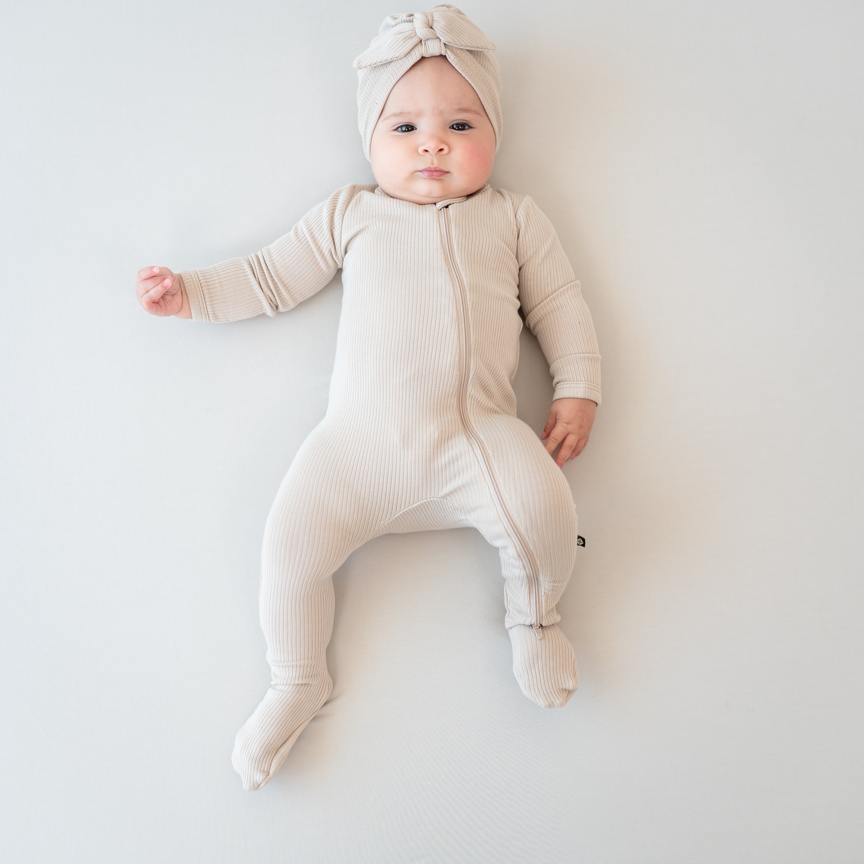 Infant girl laying down on a light neutral background wearing the Ribbed Zipper Footie in Bisque with matching ribbed headwrap