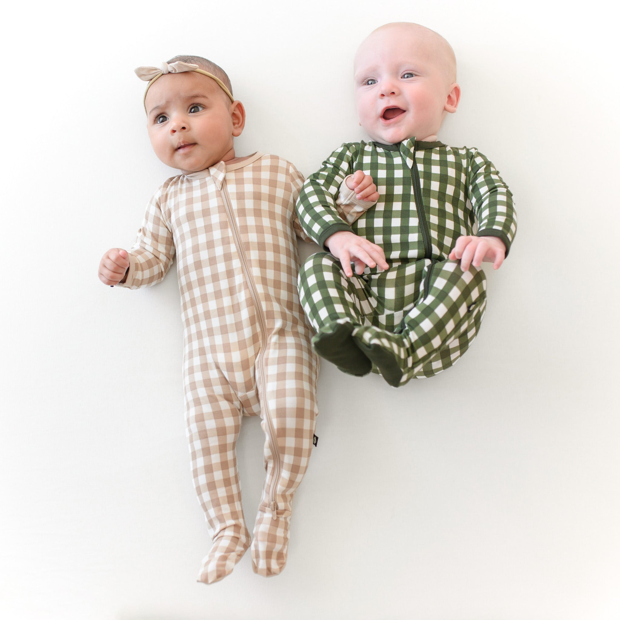 Two infant models laying side by side wearing the Zippered Footie in Gingham Fir and Gingham Bisque