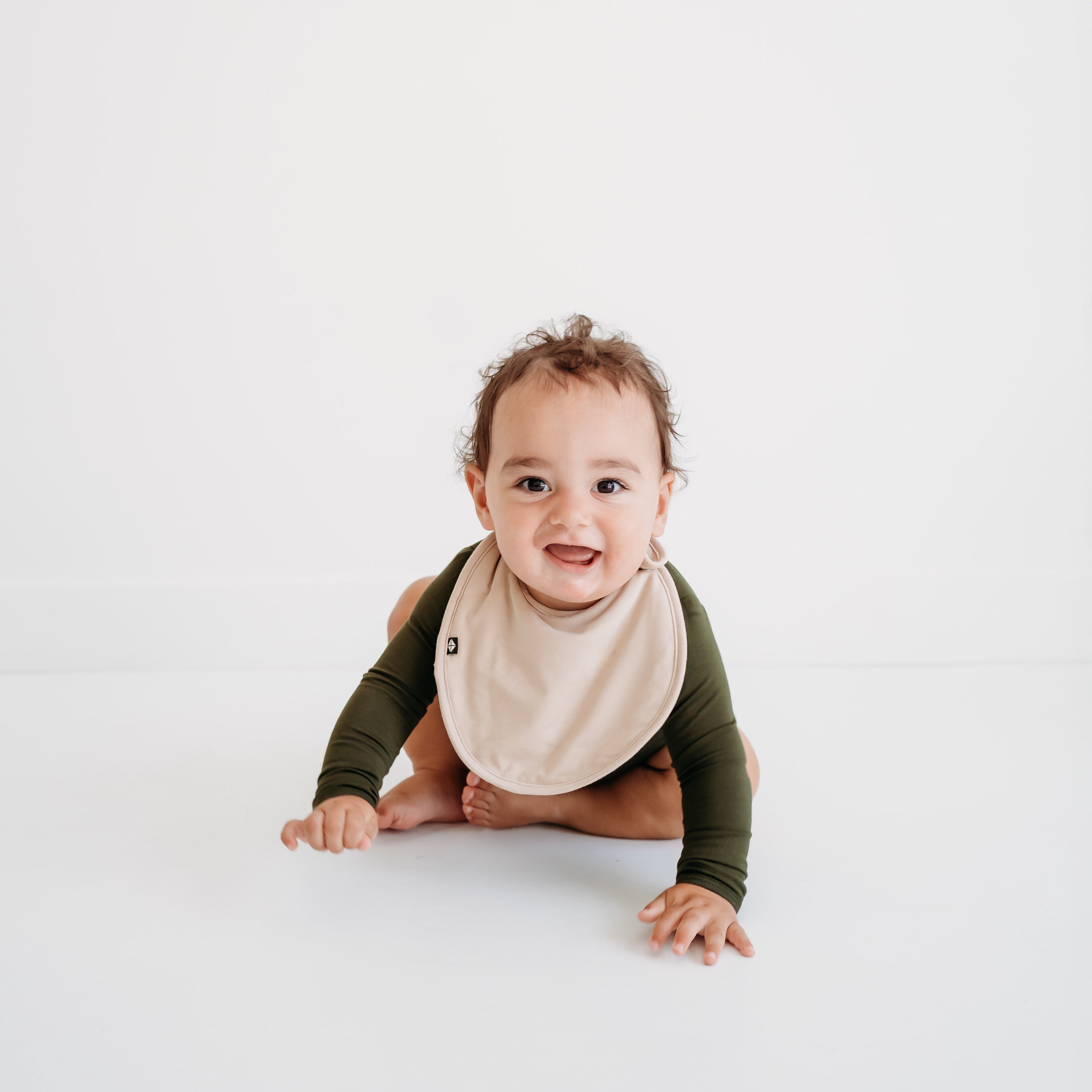Infant crawling wearing the Cushy Bib in Bisque paired with a long sleeve bodysuit in Fir