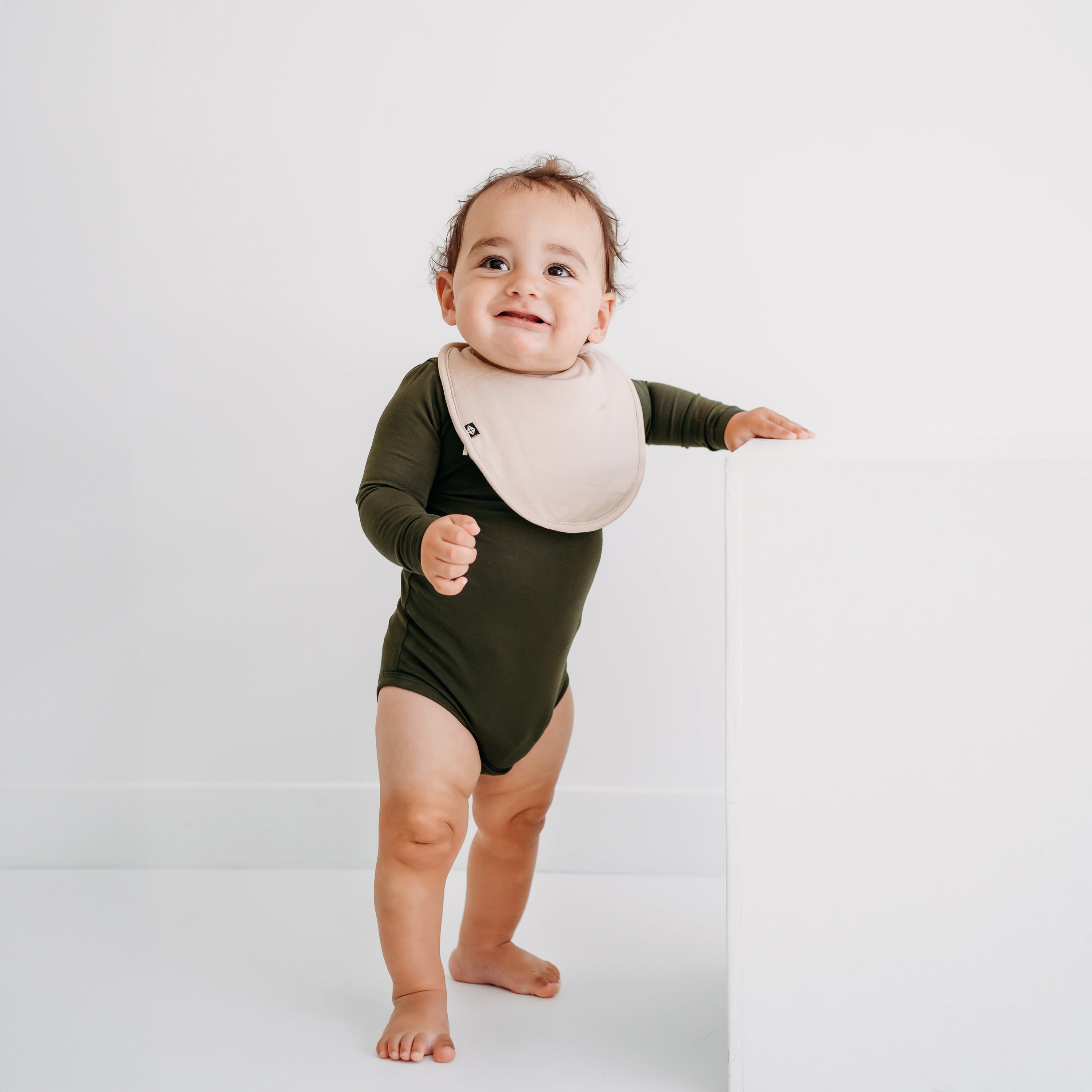 Infant standing looking up wearing the Cushy Bib in Bisque overtop a Fir long sleeve bodysuit