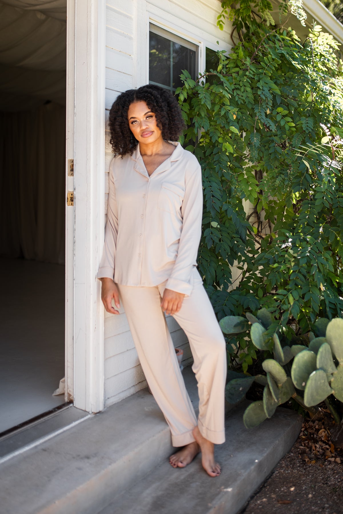 Female model leaning against a white house wearing the Long-Sleeved Women's Pajama Set in Bisque with Bisque Trim